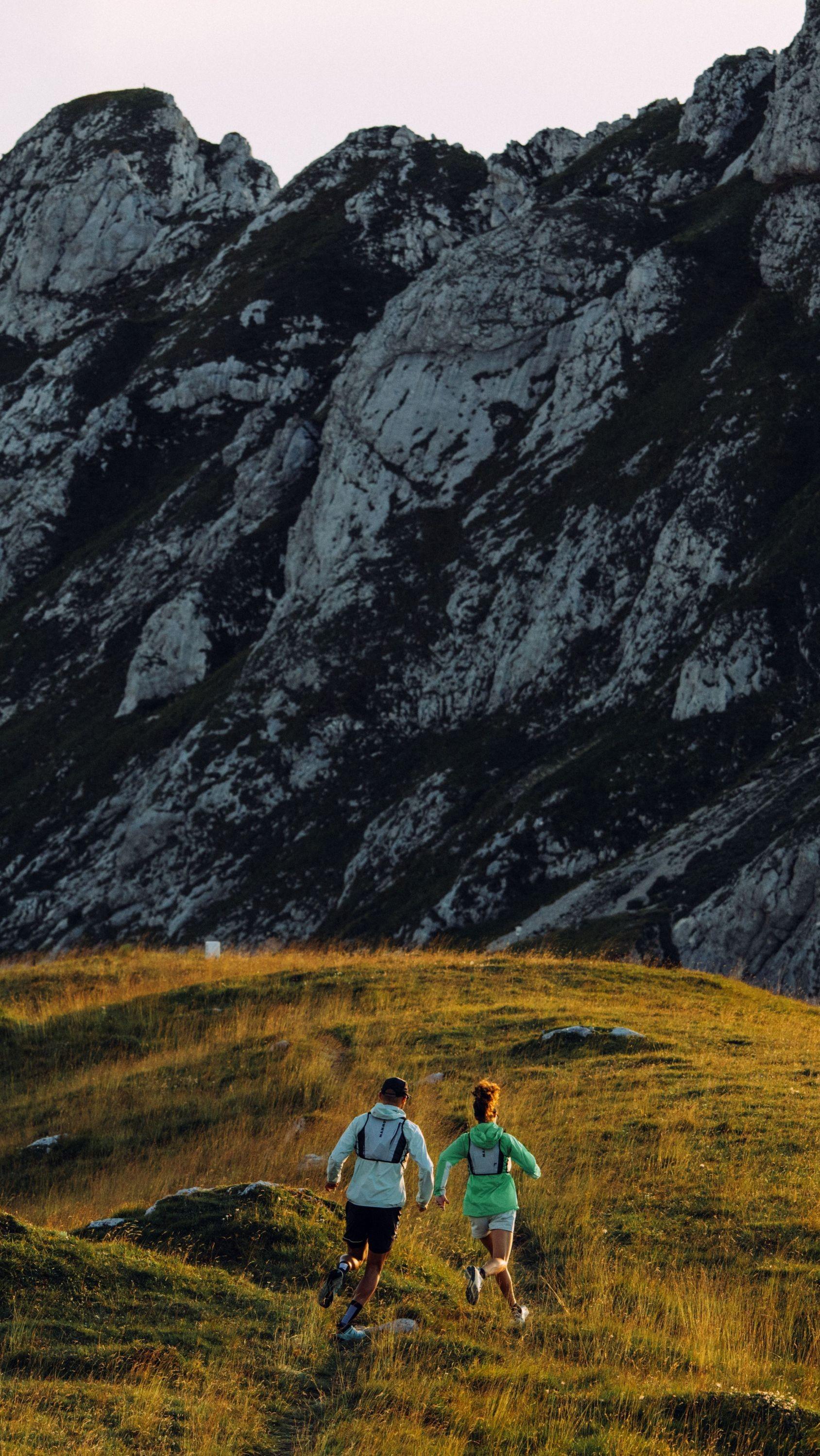 Two trail runners wearing Mammut gear run on a grassy path with rugged, rocky mountains towering in the background.
