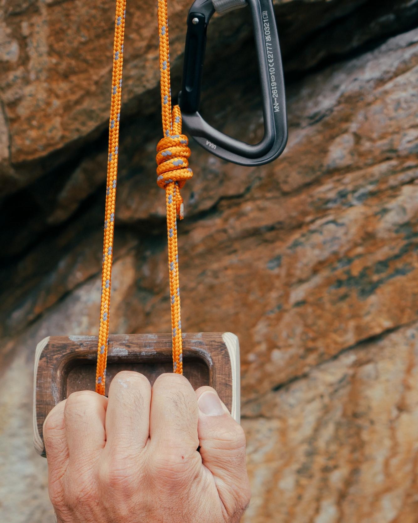 Close-up of a climber's hand gripping a wooden hold on an orange Mammut rope with a carabiner attached, set against a rocky background.