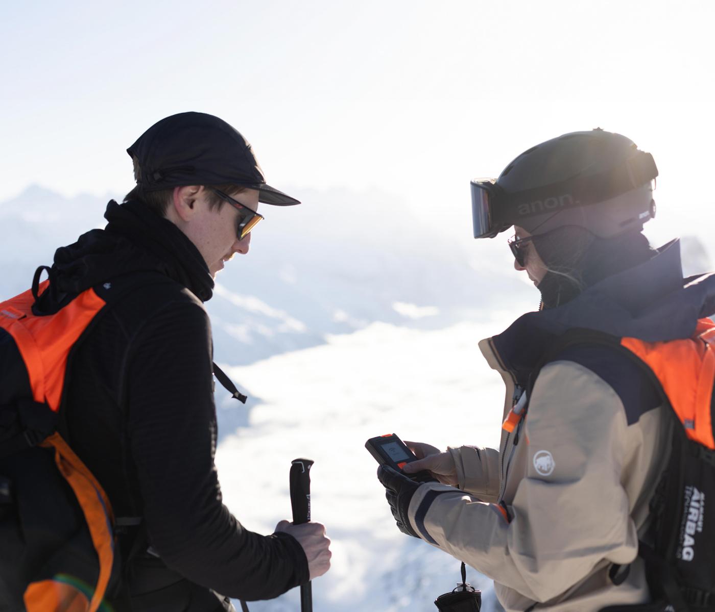Two adventurers in Mammut winter gear, equipped with backpacks, standing on a snow-covered mountain peak; one is checking their phone.
