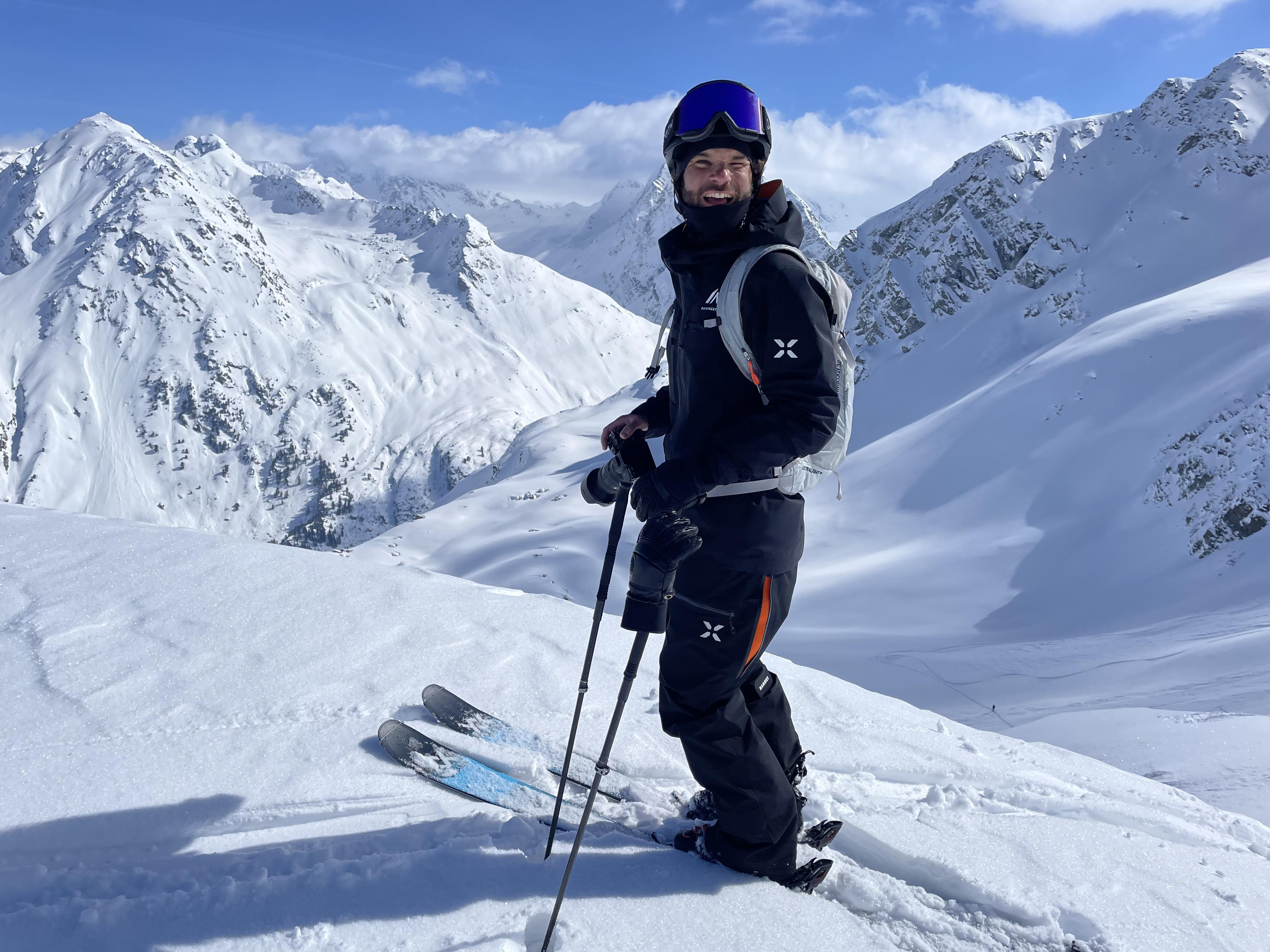 A skier wearing Mammut gear stands on snowy mountain slopes, smiling with ski poles in hand, surrounded by impressive snow-covered alpine peaks.