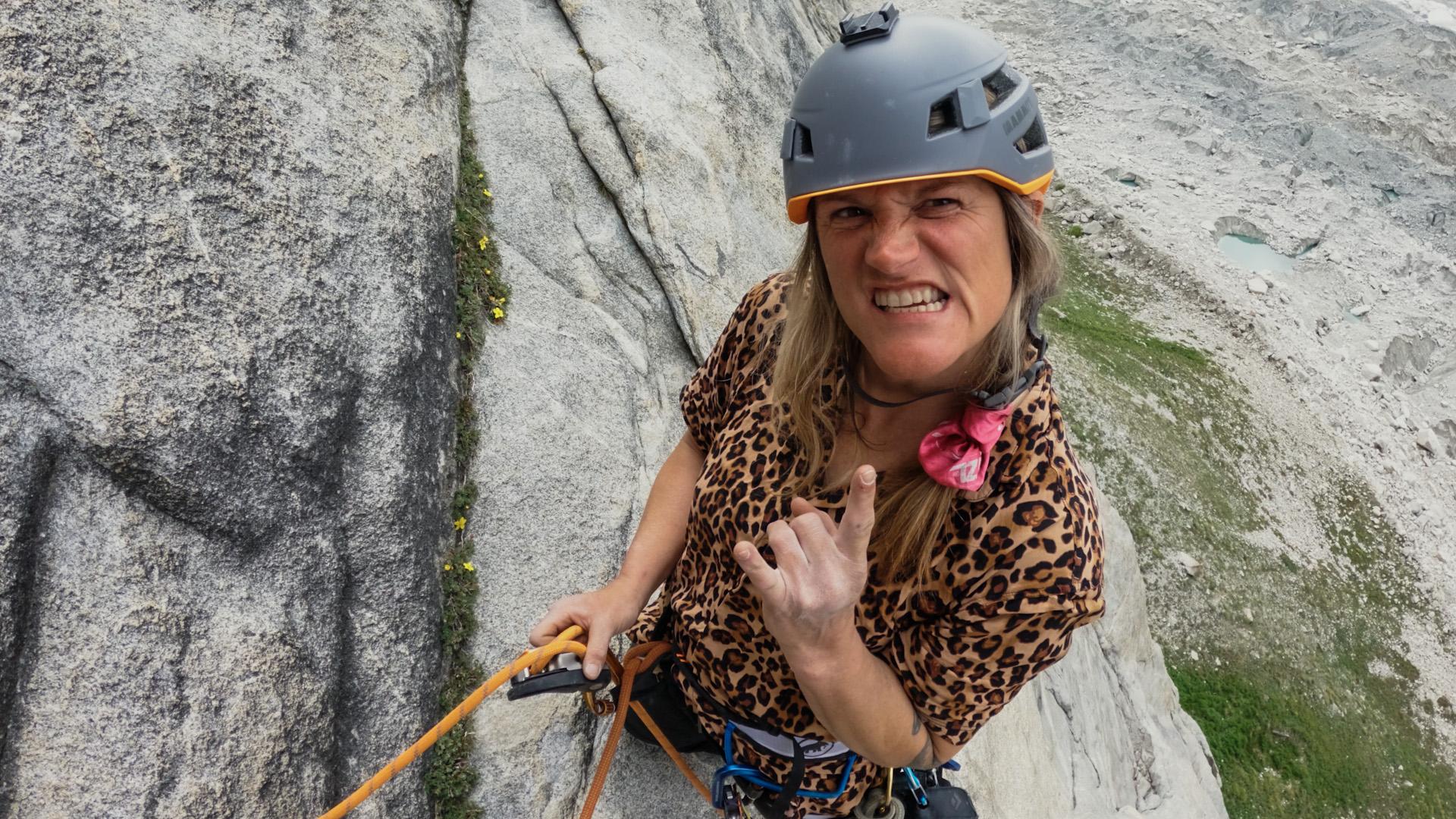 A climber in a leopard print shirt and helmet, wearing Mammut climbing gear, smiles and makes a hand gesture while scaling a rocky cliff.