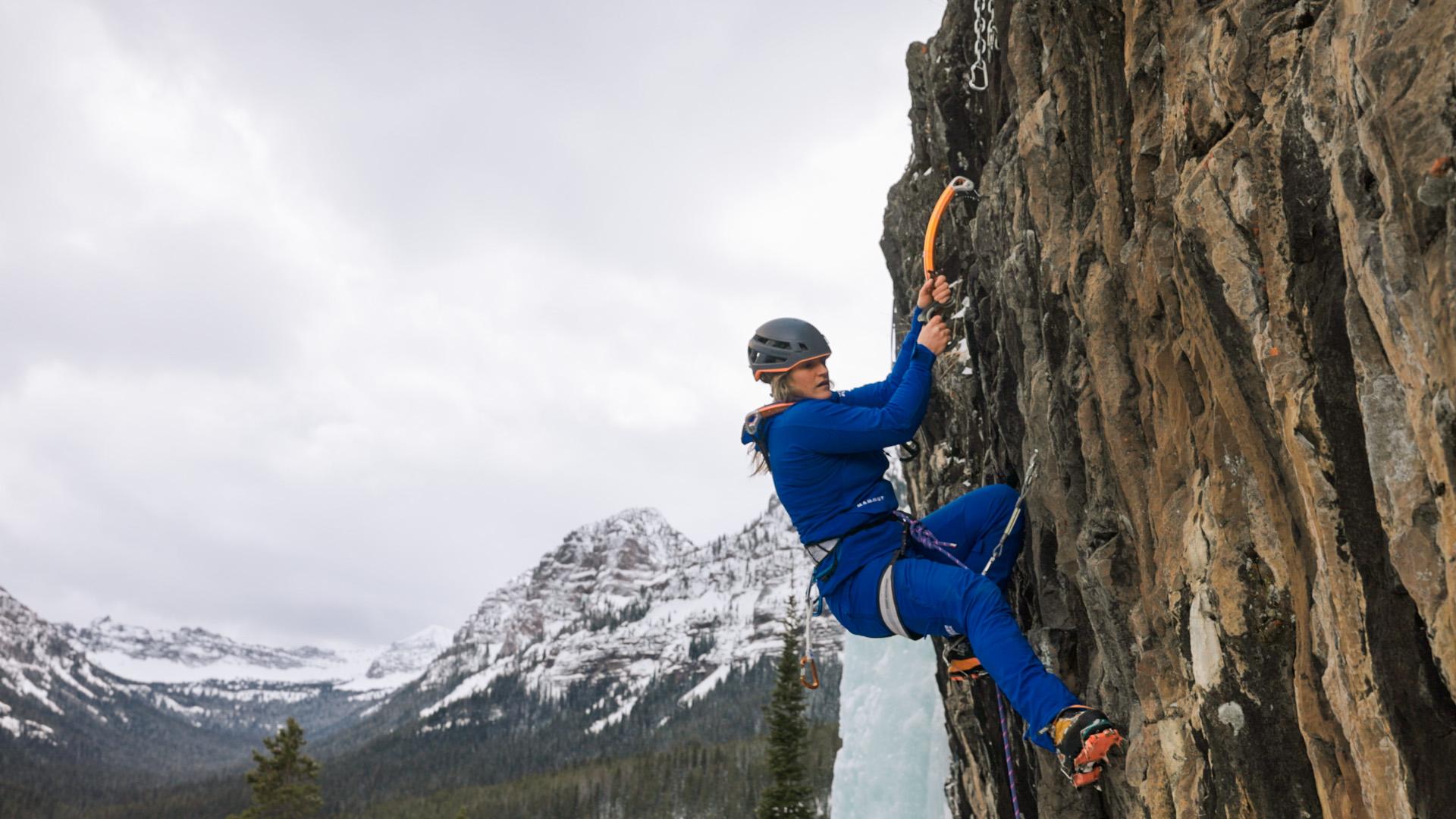 Mammut-clad climber in blue gear ice climbing a rocky cliff, with snowy alpine mountains in the background.