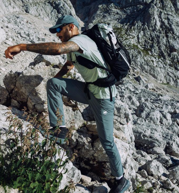 A man wearing Mammut hiking gear climbs rocky terrain with a backpack in an alpine mountain landscape.