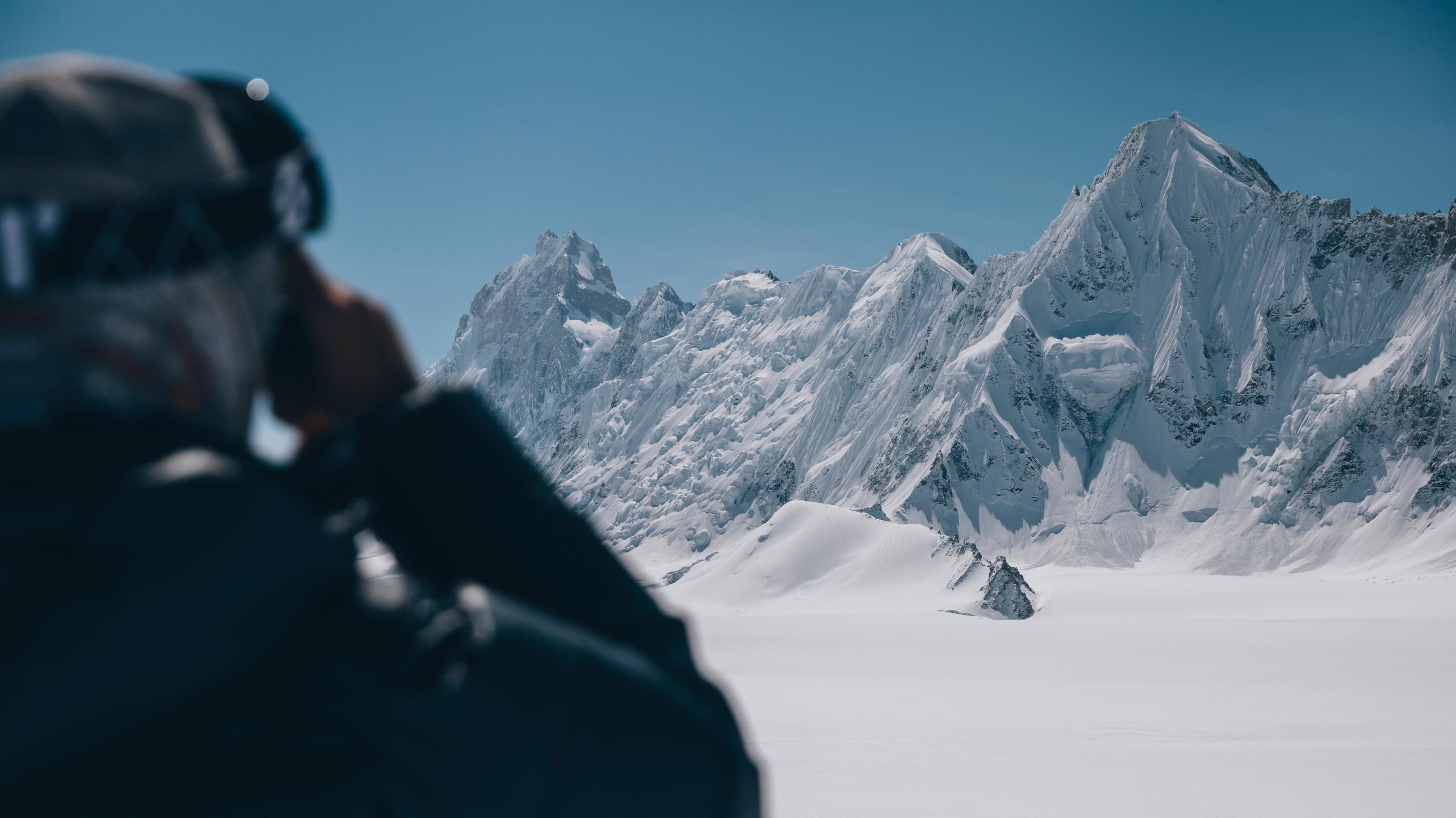 A skier outfitted in Mammut gear gazes at snow-covered mountains under a clear blue sky.