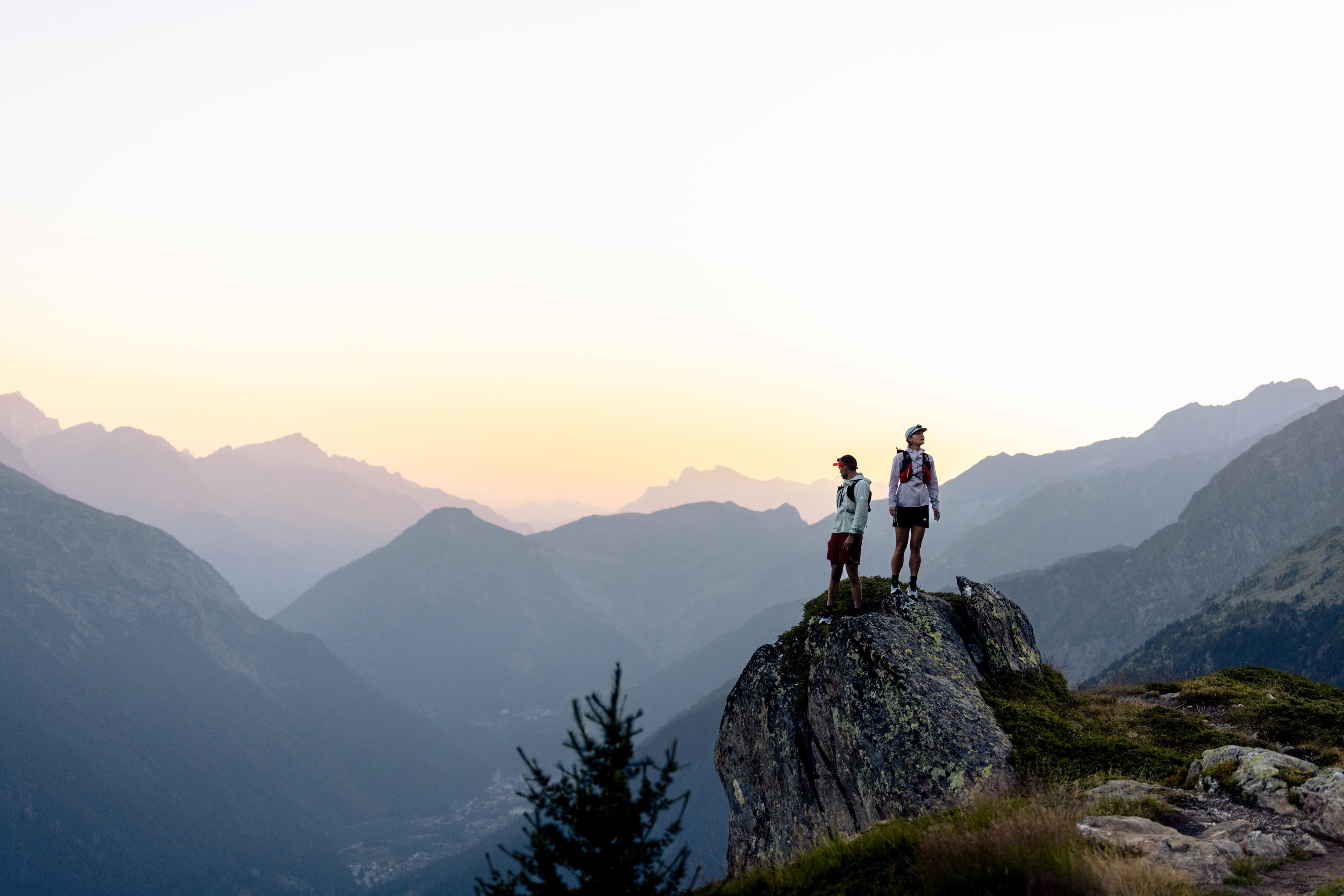 Three hikers in Mammut outdoor gear ascend a rocky mountain trail, surrounded by misty peaks and sunlight in the background.