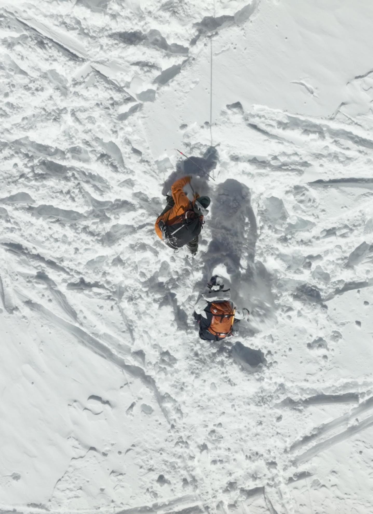Two adventurers in vibrant Mammut jackets navigate snowy terrain, casting long shadows on the snow, as seen from above.
