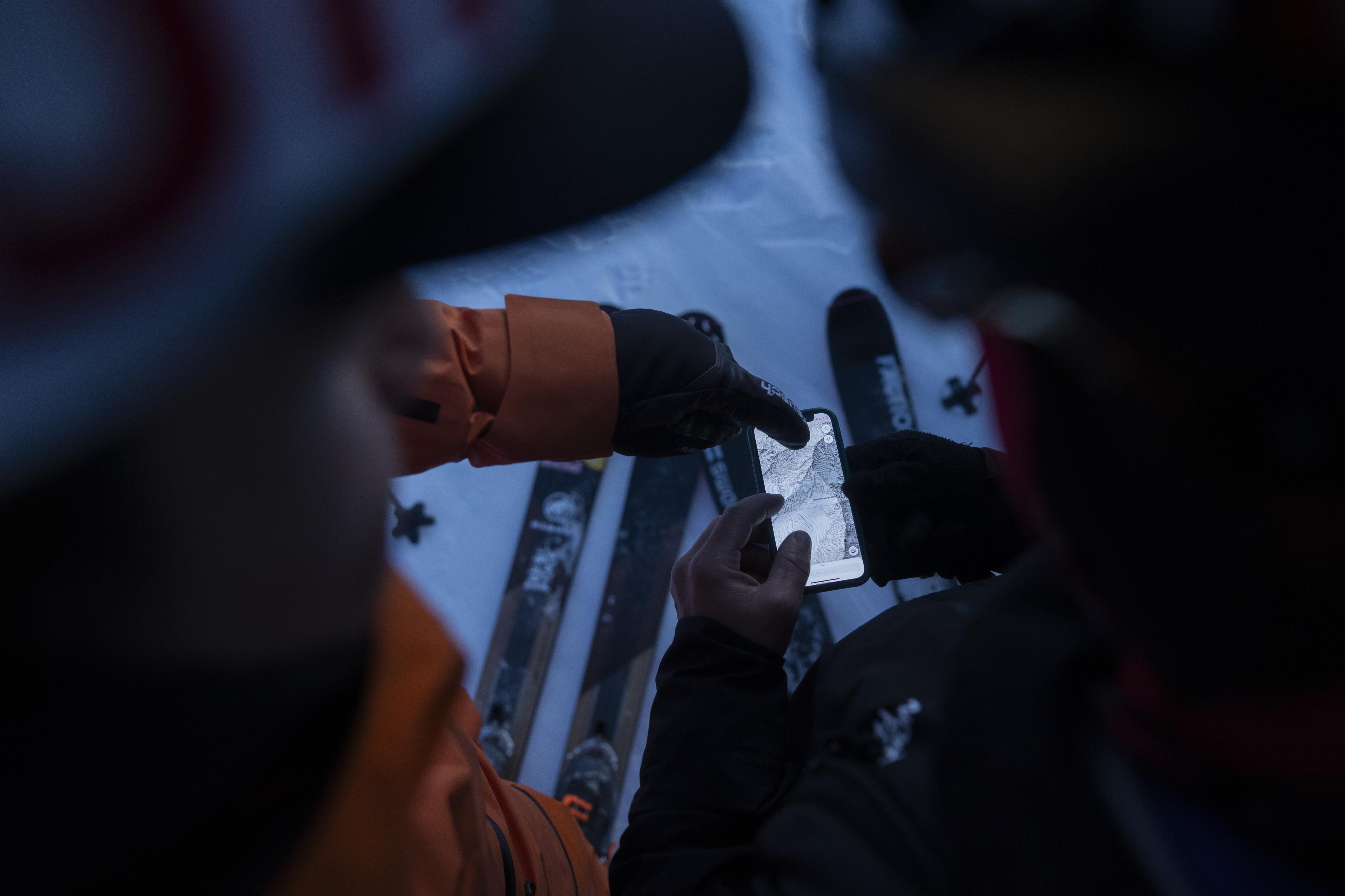 Two skiers check a map or phone screen while sitting on snowy ground, surrounded by Mammut skis and gear.