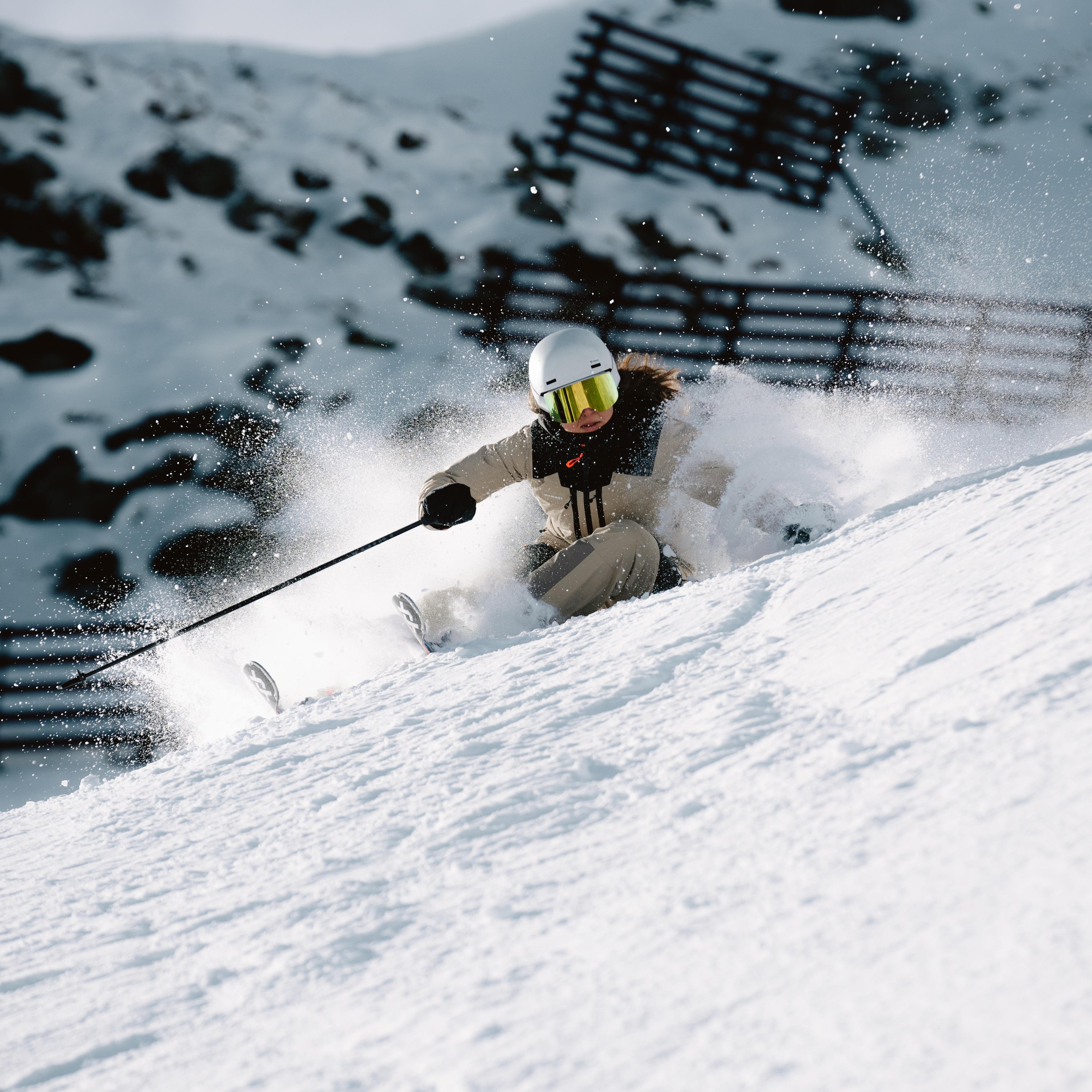 Anne Kissling, a skier, in a helmet and goggles expertly navigates through fresh powder on a sunny mountain slope, showcasing Mammut's premium mountaineering gear.