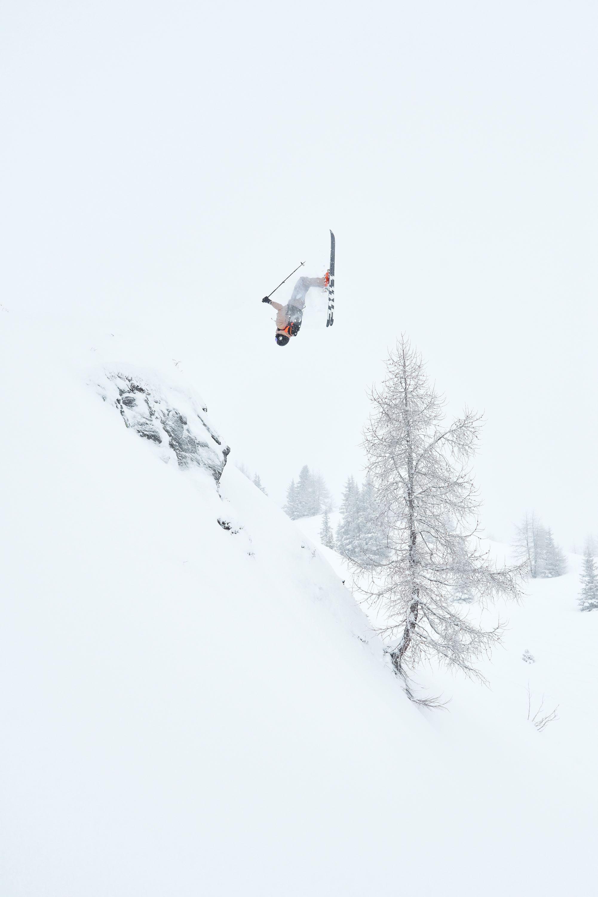 Skier in Mammut gear performing a backflip in mid-air against a snowy backdrop with a tree and snow-covered slope.