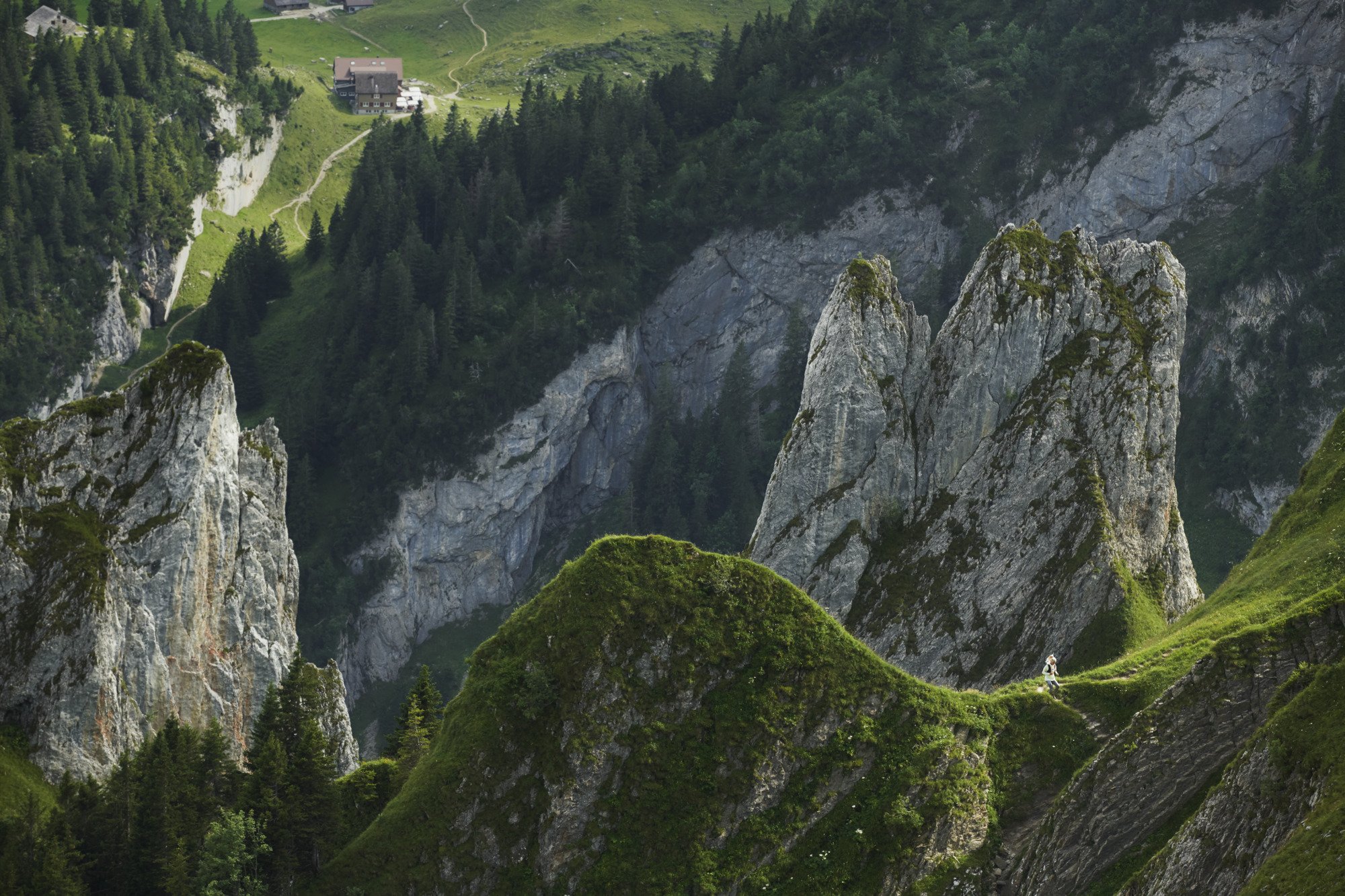 Dramatic mountain peaks covered in lush green grass, perfect for Mammut mountaineering. A distant house nestled among trees adds a picturesque touch to the pristine landscape.
