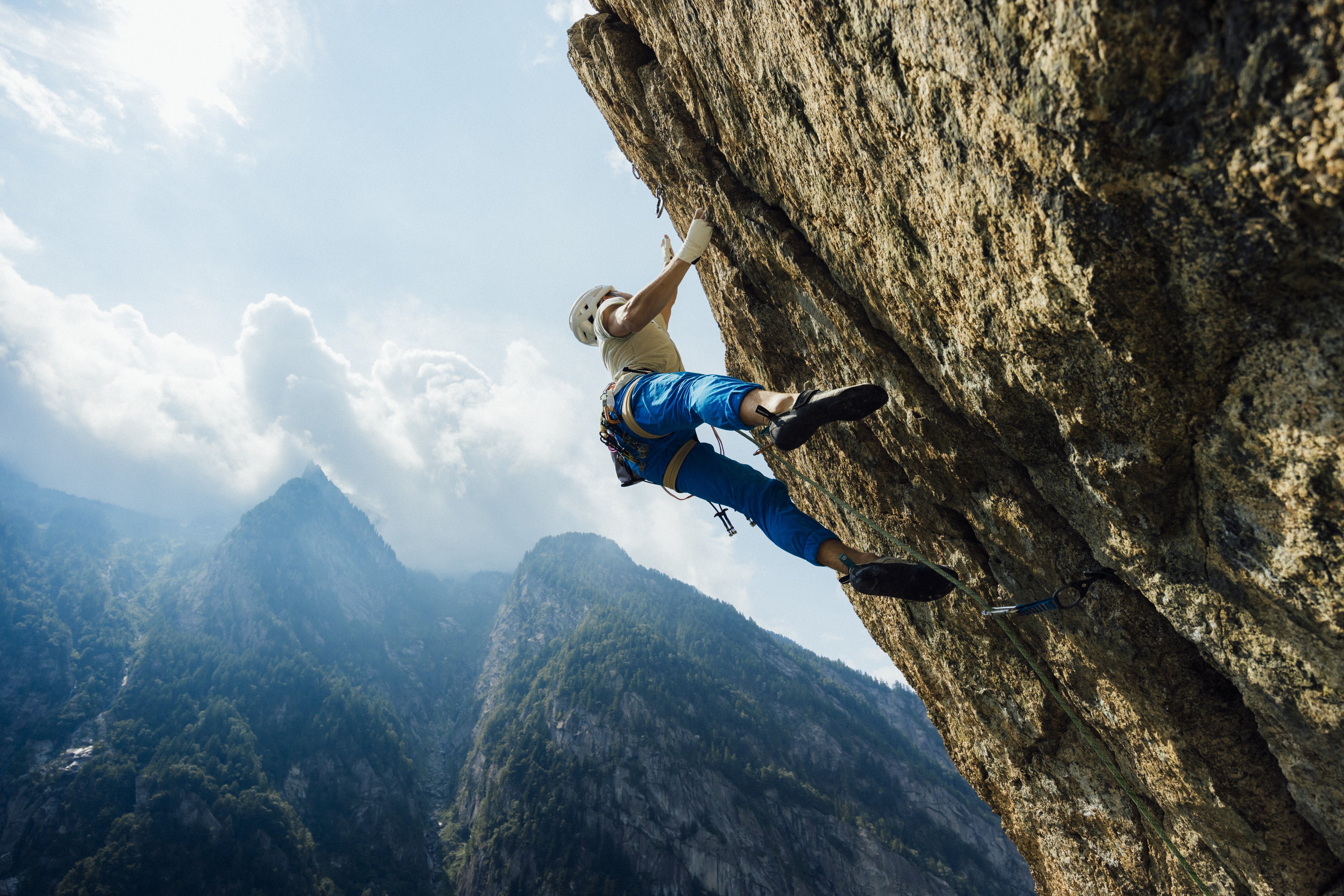 A climber wearing blue pants ascends a rugged rock face in the mountains, equipped with Mammut climbing gear, with dramatic jagged peaks and a cloudy sky in the background.