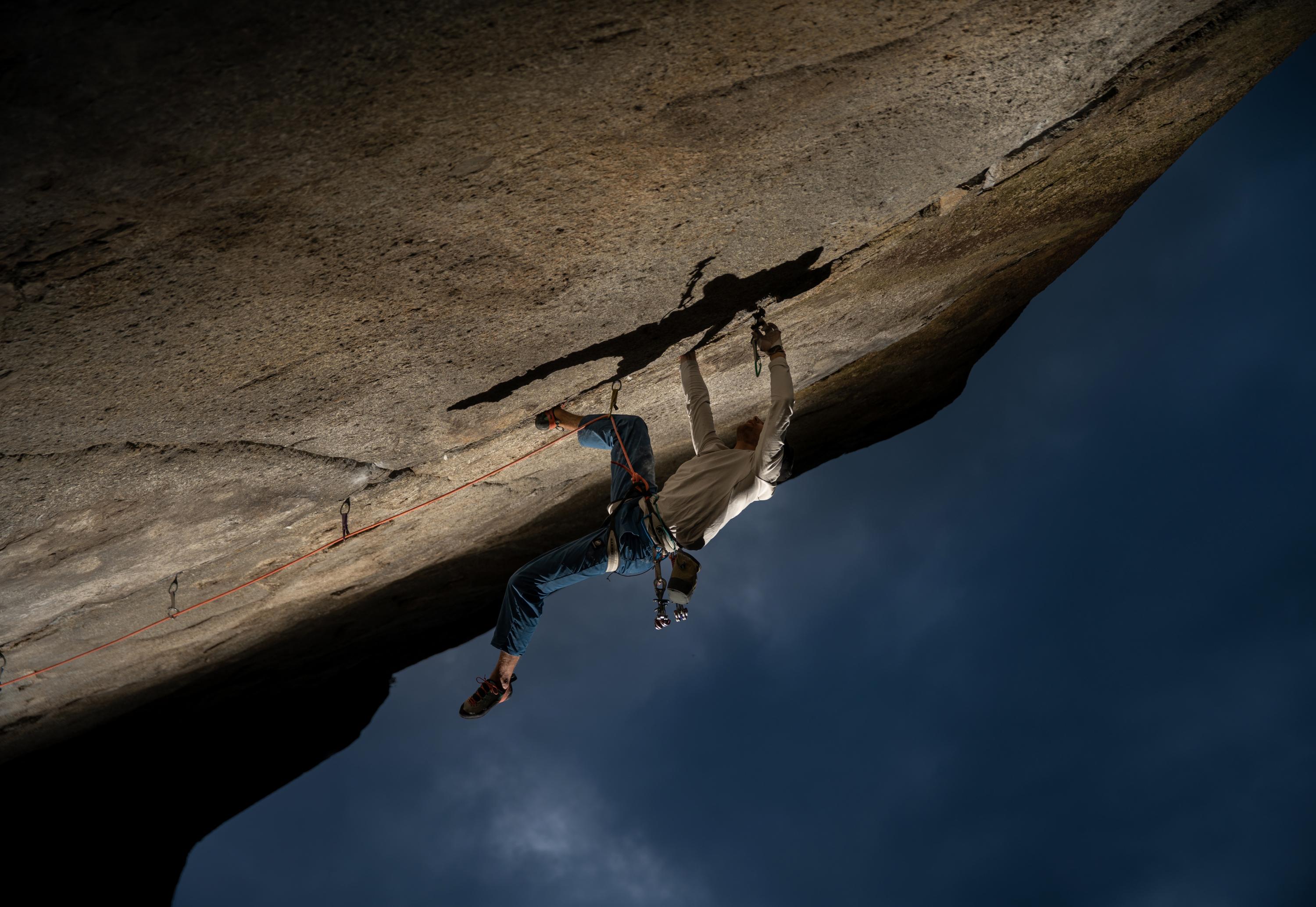 A climber wearing a white shirt and blue pants, equipped with Mammut gear, scales an overhanging rock face under a dark sky.