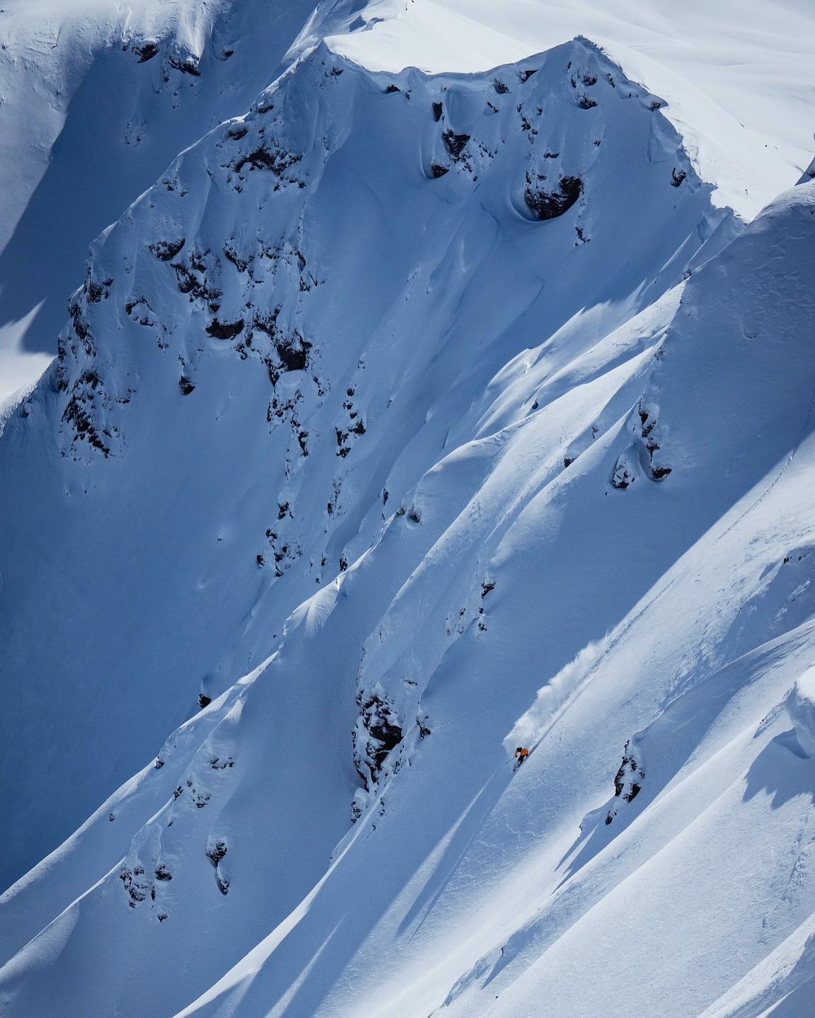 A lone skier in Mammut gear descends a steep, snowy mountain slope surrounded by rugged, snow-covered cliffs.