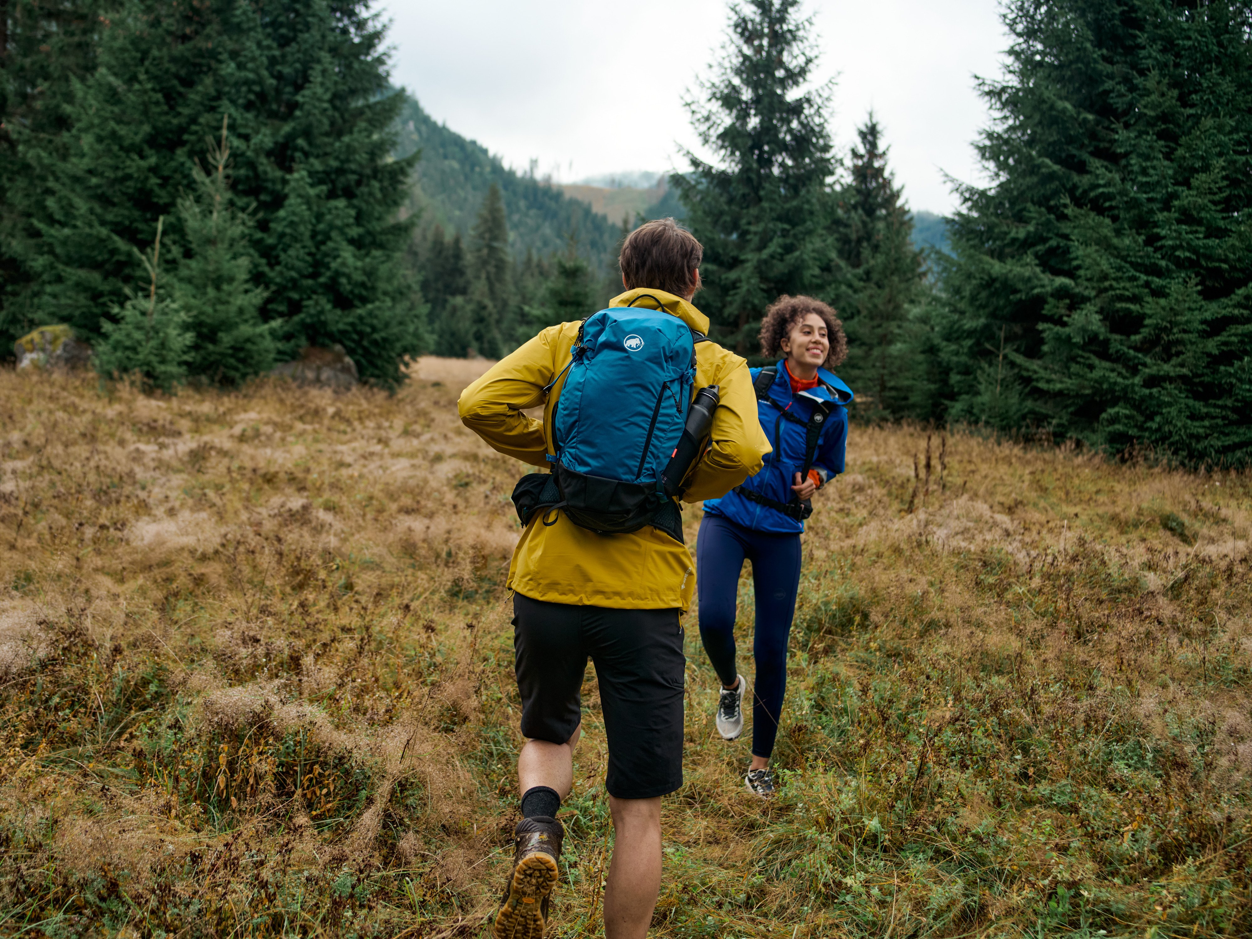Two hikers wearing Mammut backpacks, laughing as they trek through a grassy, forested mountain area.