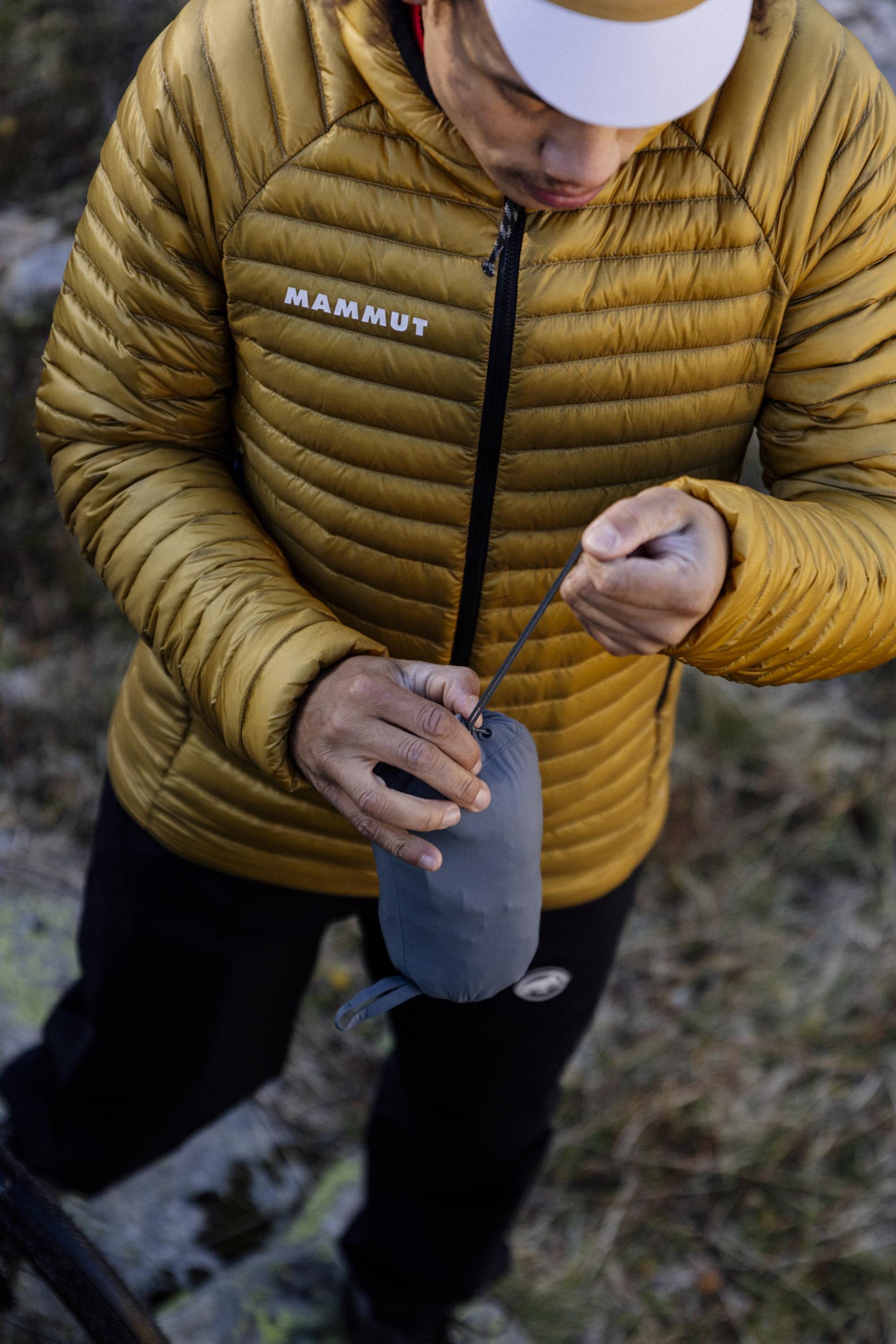 Person wearing a yellow Mammut jacket and cap compressing a gray outdoor gear bag.