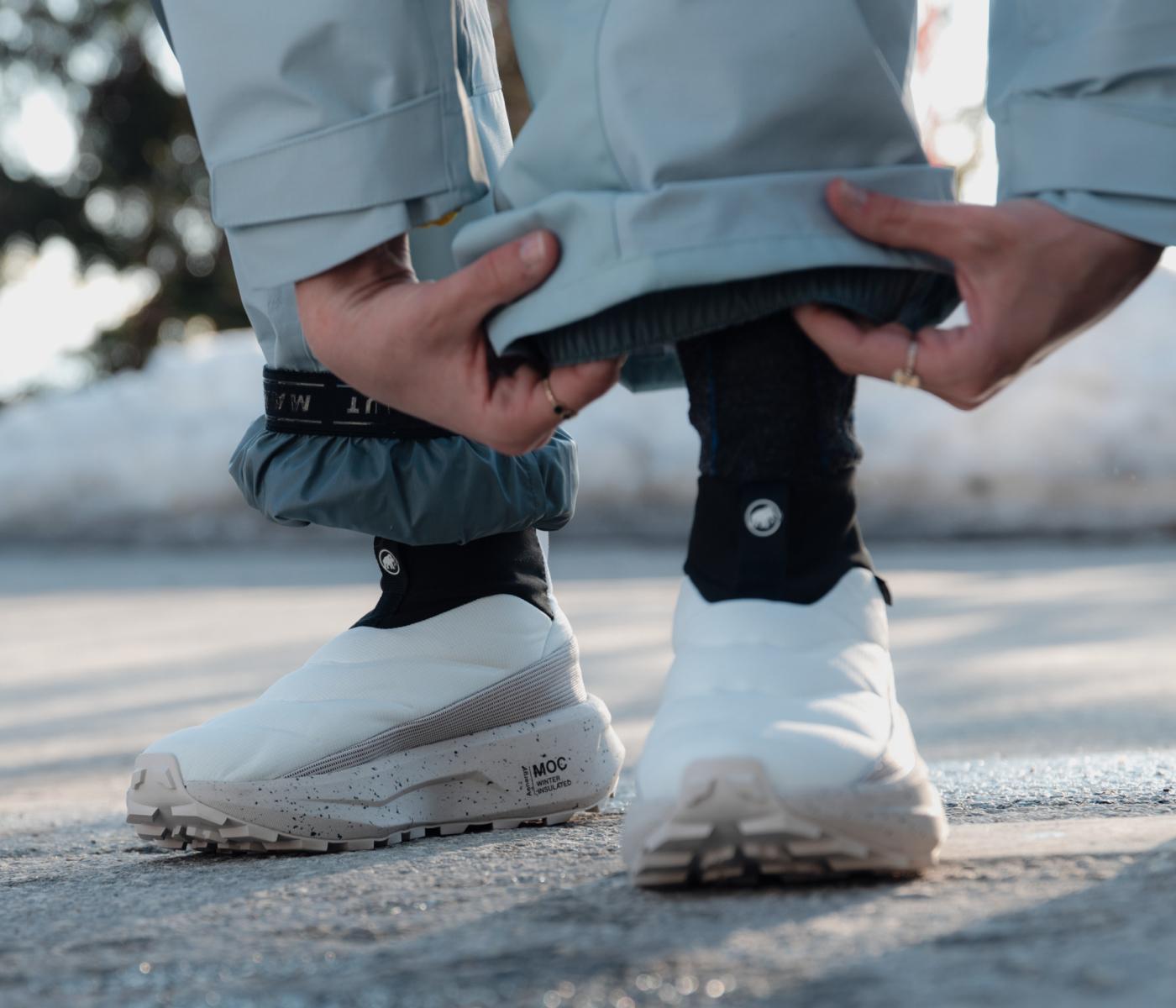 Close-up of a person adjusting pant leg over white Mammut outdoor shoes with black gaiters, standing on pavement.