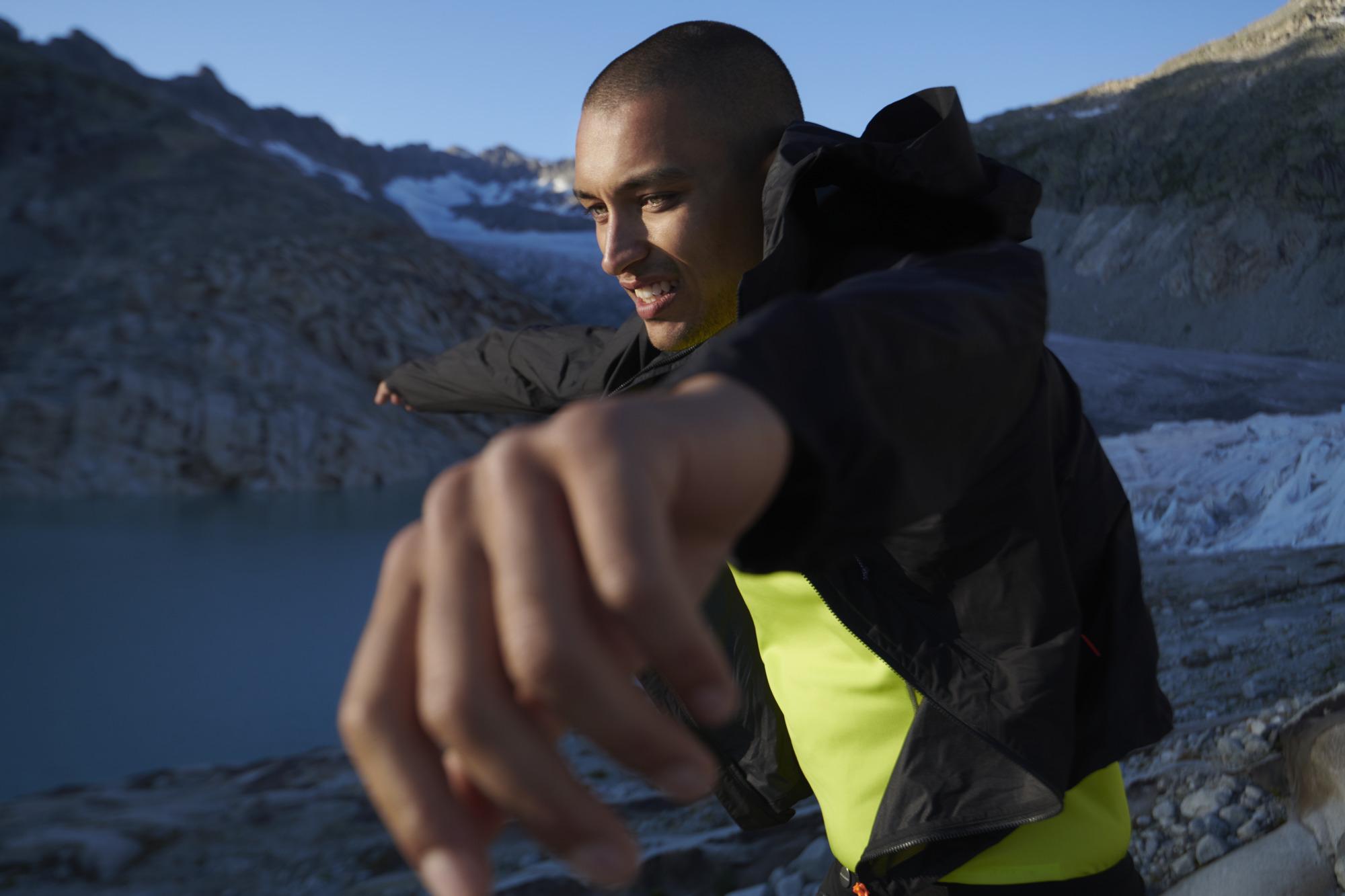 Person in Mammut athletic wear stretches against a rugged mountainous backdrop near an alpine lake.