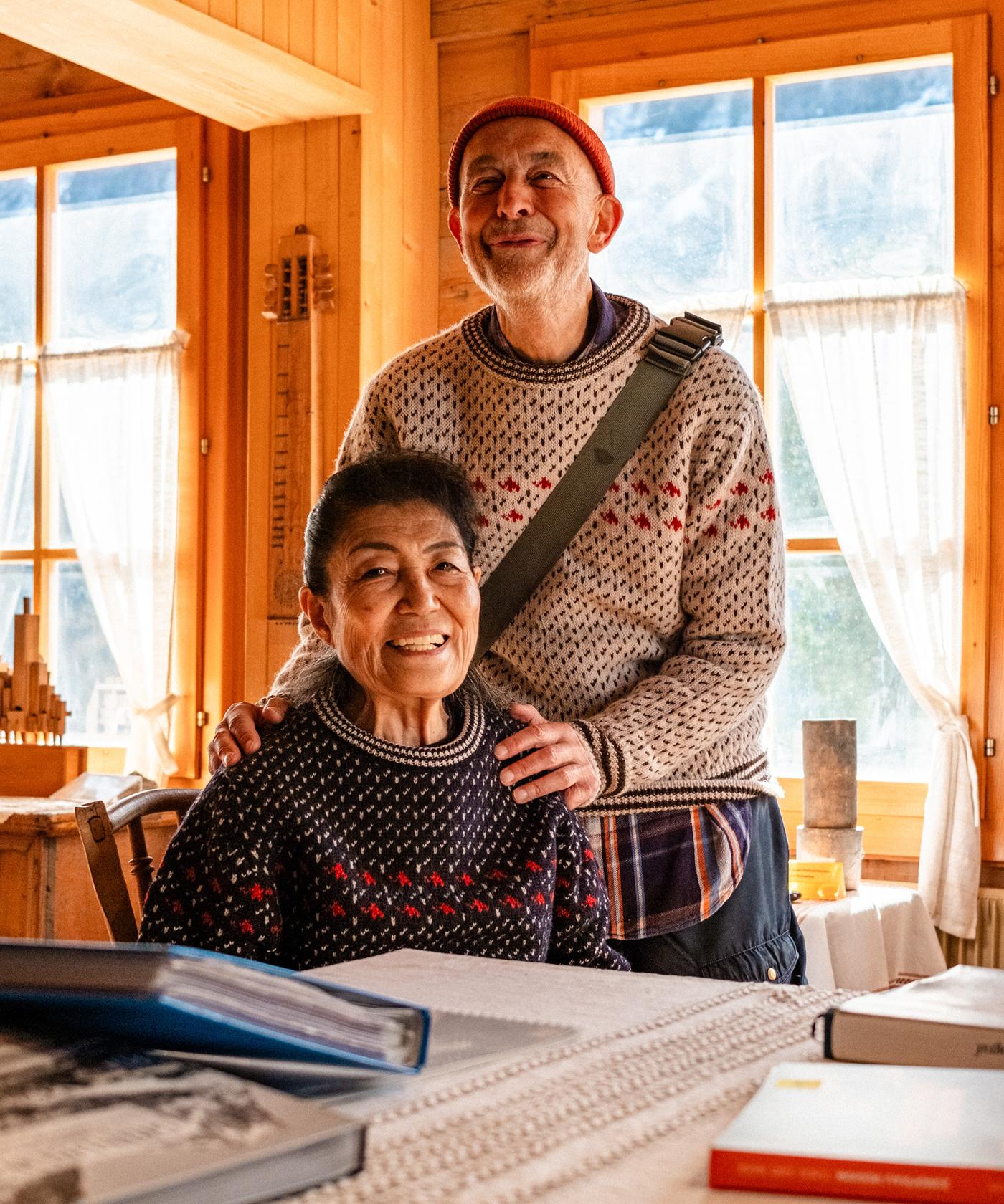 Two smiling people in Mammut sweaters pose together in a sunlit wooden room with books on the table, highlighting cozy outdoor style.