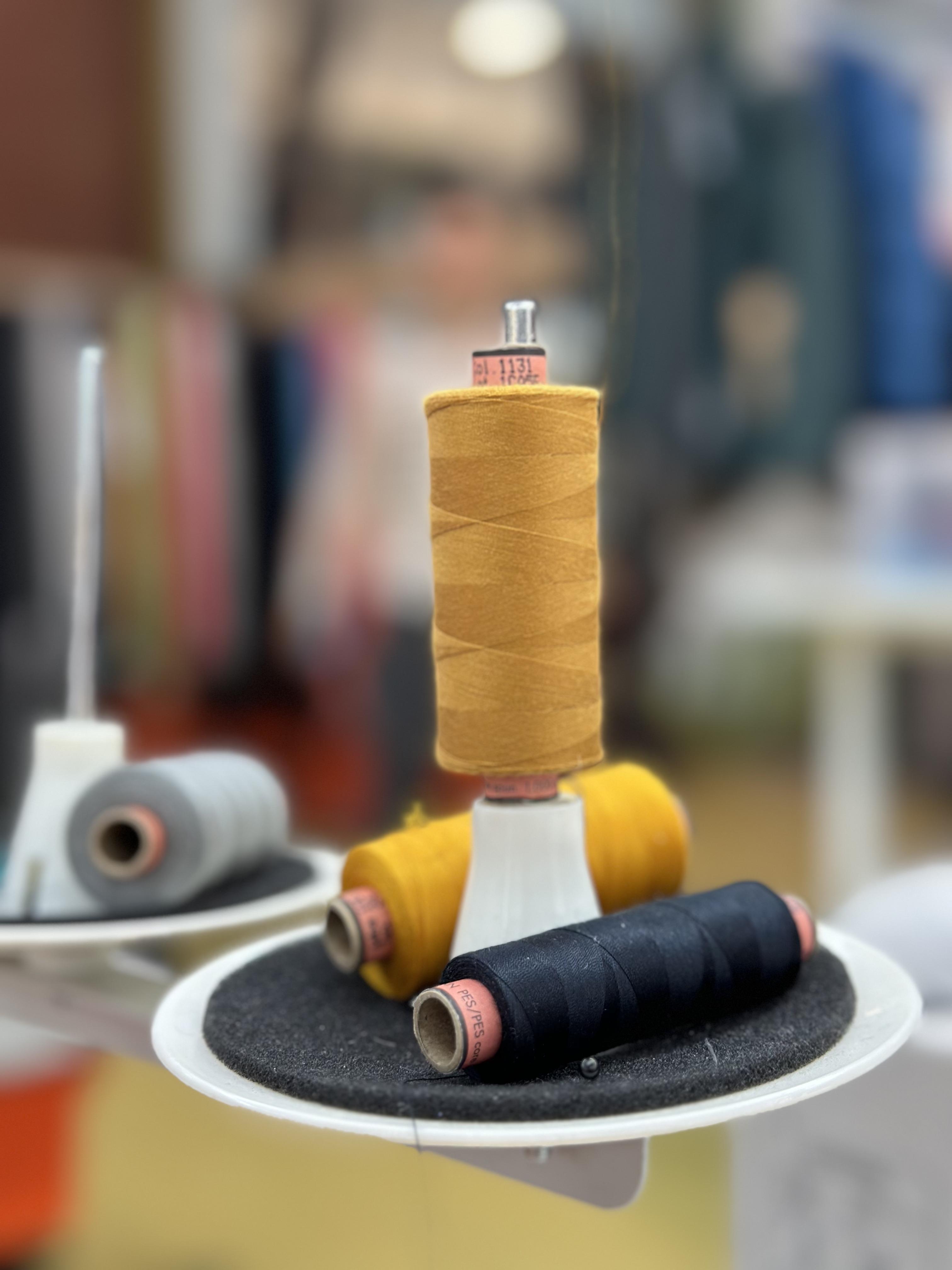 Close-up of yellow and black thread spools on a white sewing machine attachment, highlighting Mammut's precision in creating high-quality sports and mountaineering gear, set against a blurred background workspace.