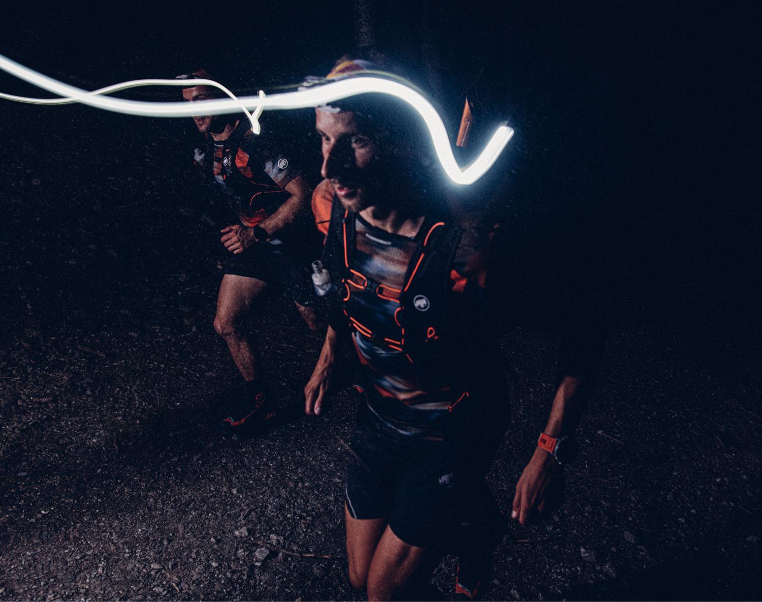Two night trail runners with headlamps, one leaving a light trail, running on a gravel path in a dimly lit forest, equipped with Mammut gear.