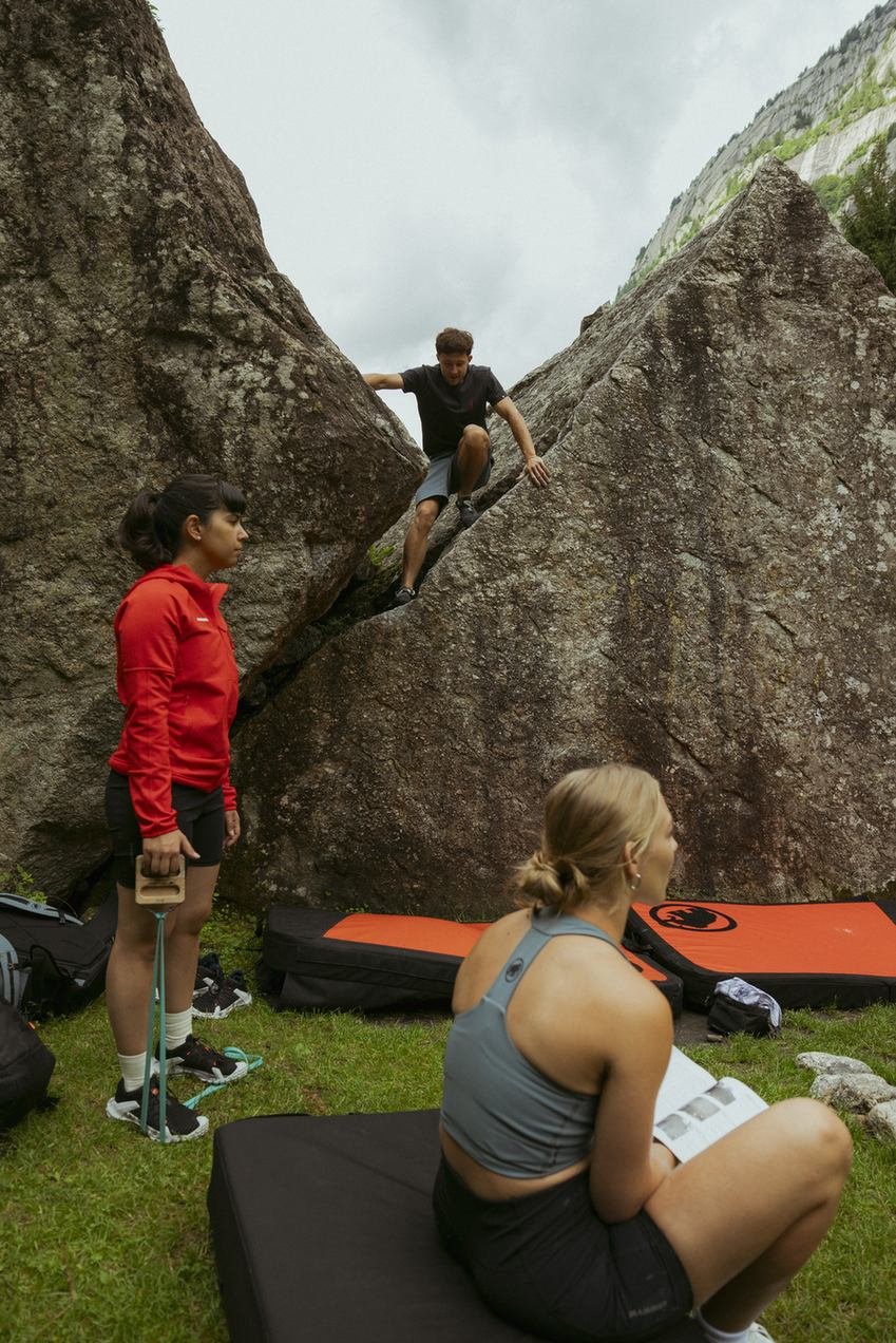 Smiling climber sits on a crash pad outdoors, showing chalked and taped hands, with Mammut climbing gear visible.
