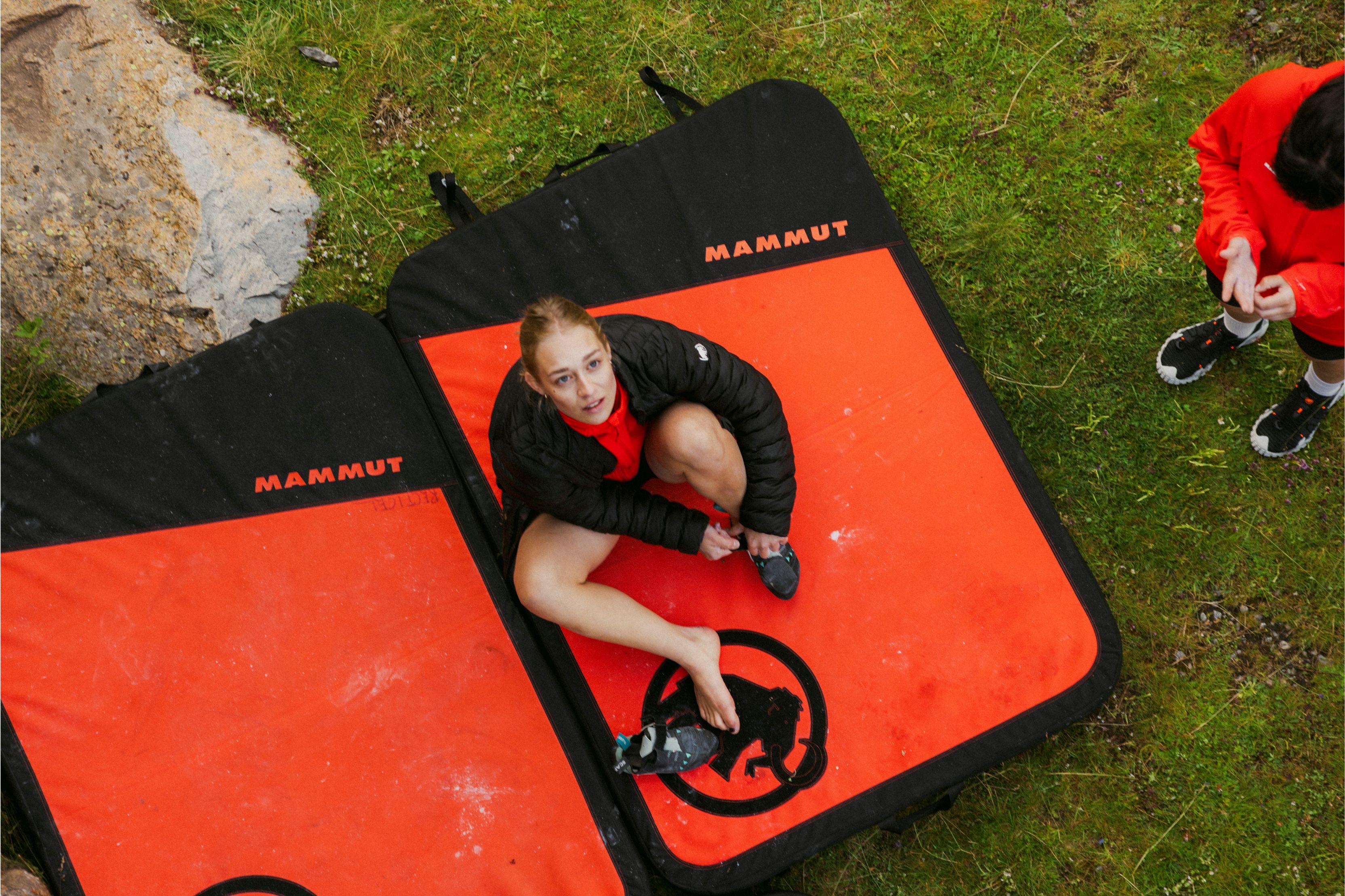 A woman sits on a Mammut crash pad outdoors, putting on climbing shoes, viewed from above, preparing for bouldering.