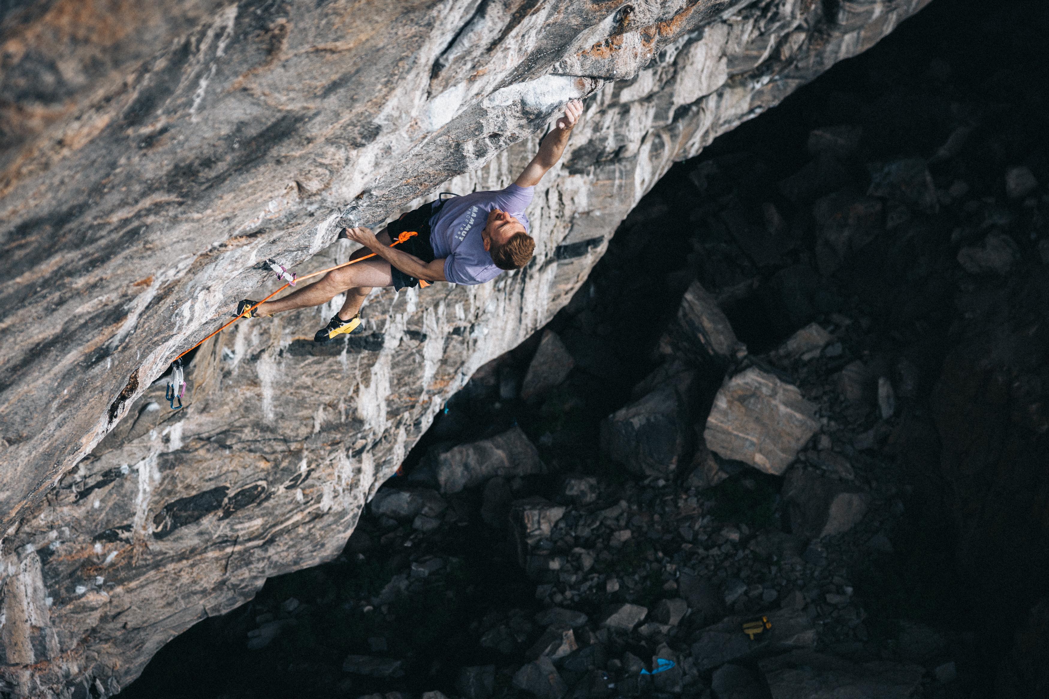 A climber wearing Mammut gear grips a steep rock face, secured by a rope, as seen from above.