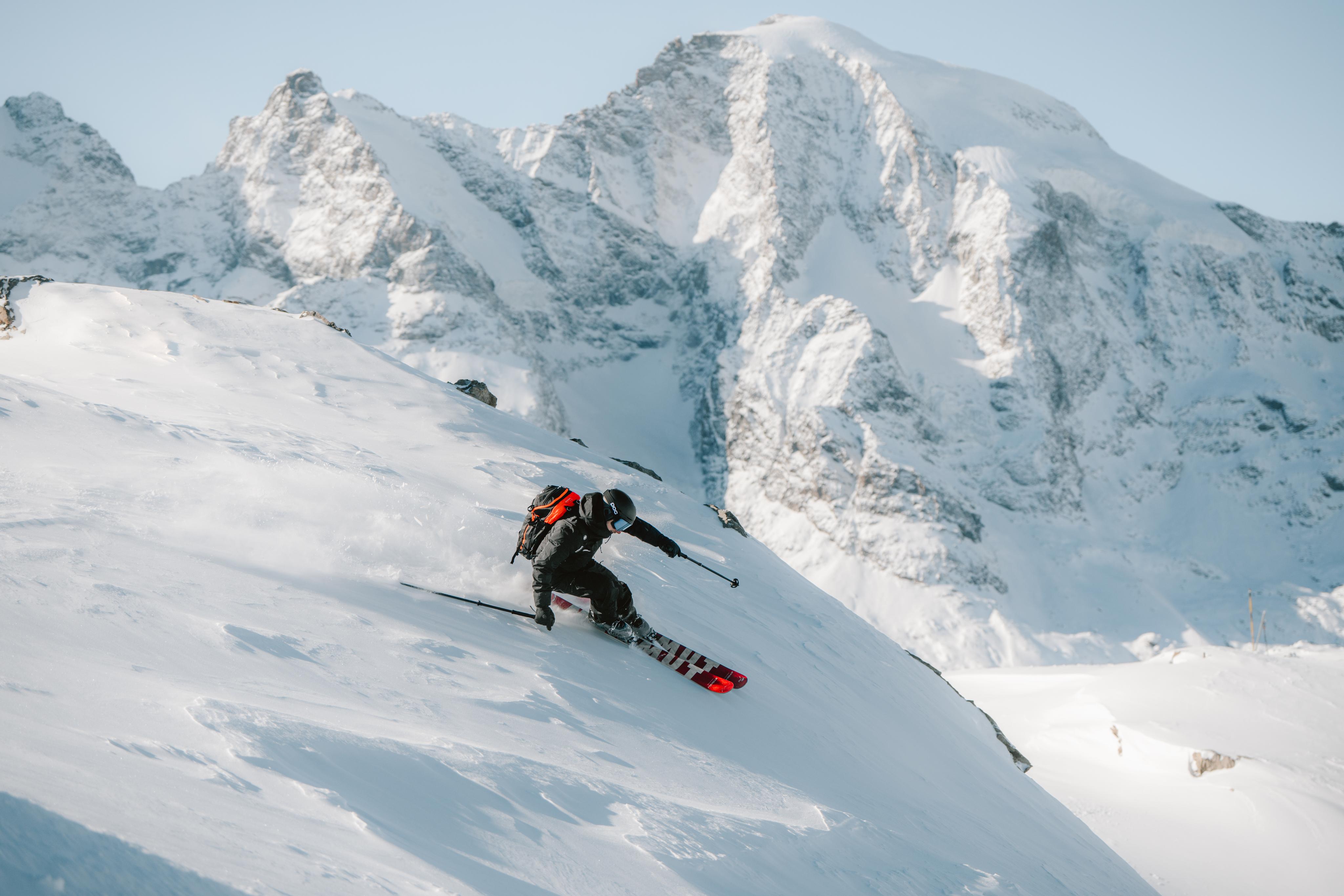 Person in pink Mammut ski gear and goggles carries skis on shoulder against a snowy mountain background.