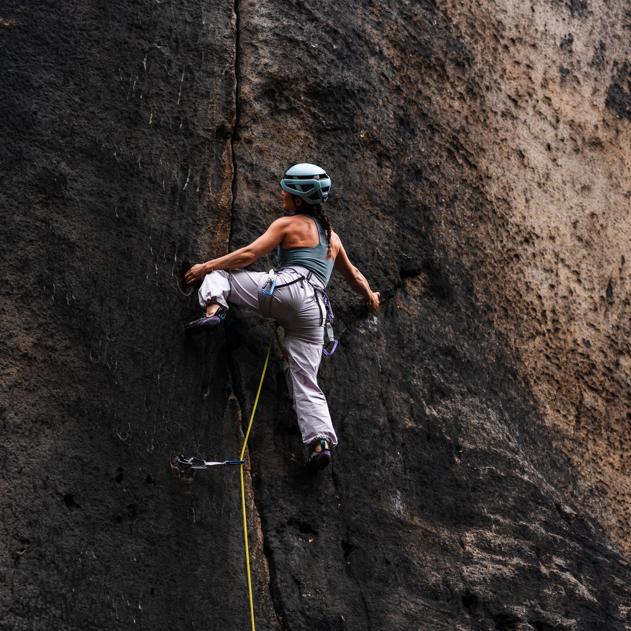 A climber equipped with a helmet and Mammut gear skillfully ascends a rock face, utilizing a crack for grip.