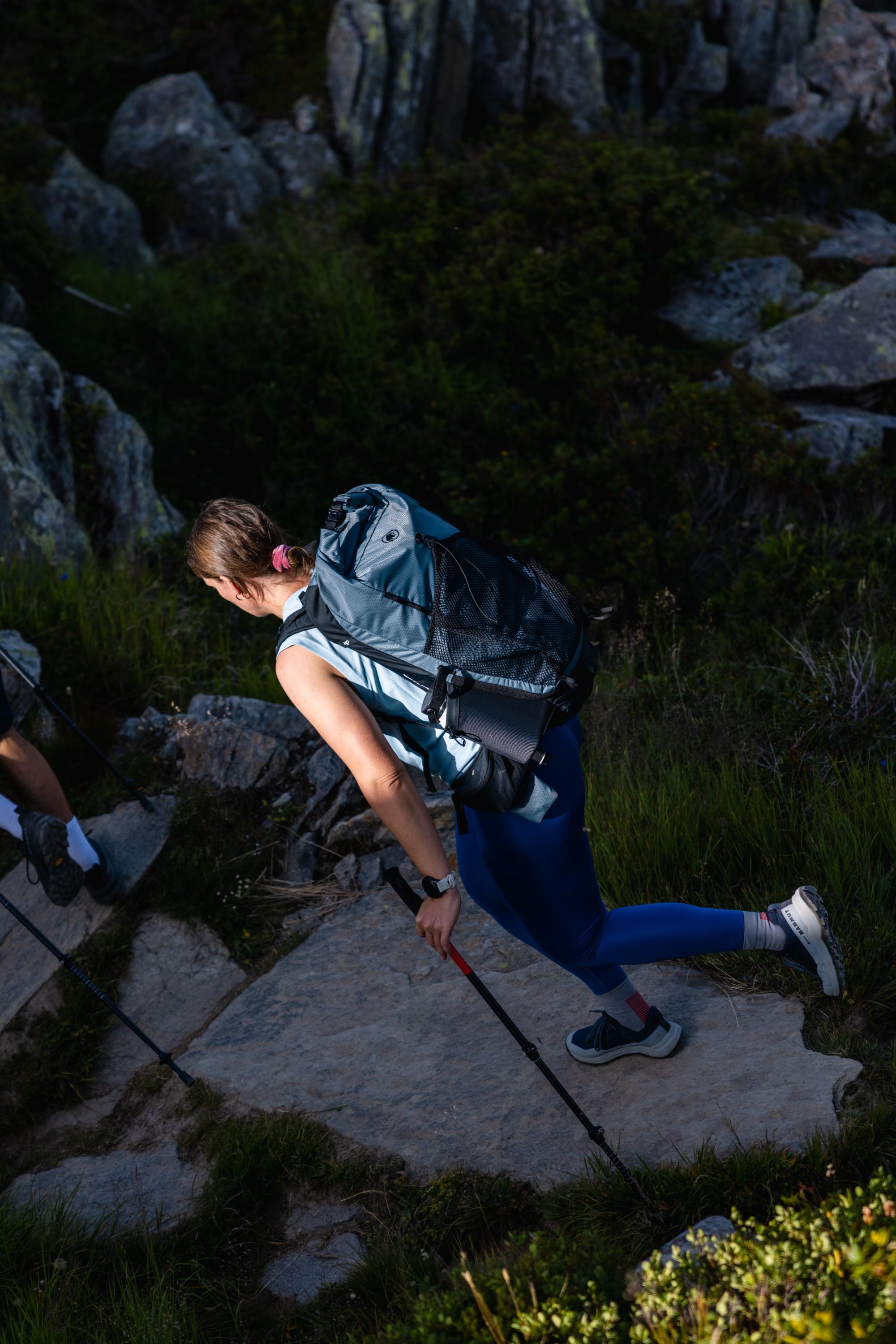 Woman wearing Mammut gear with a backpack, hiking up a rocky mountain trail surrounded by lush greenery and boulders.