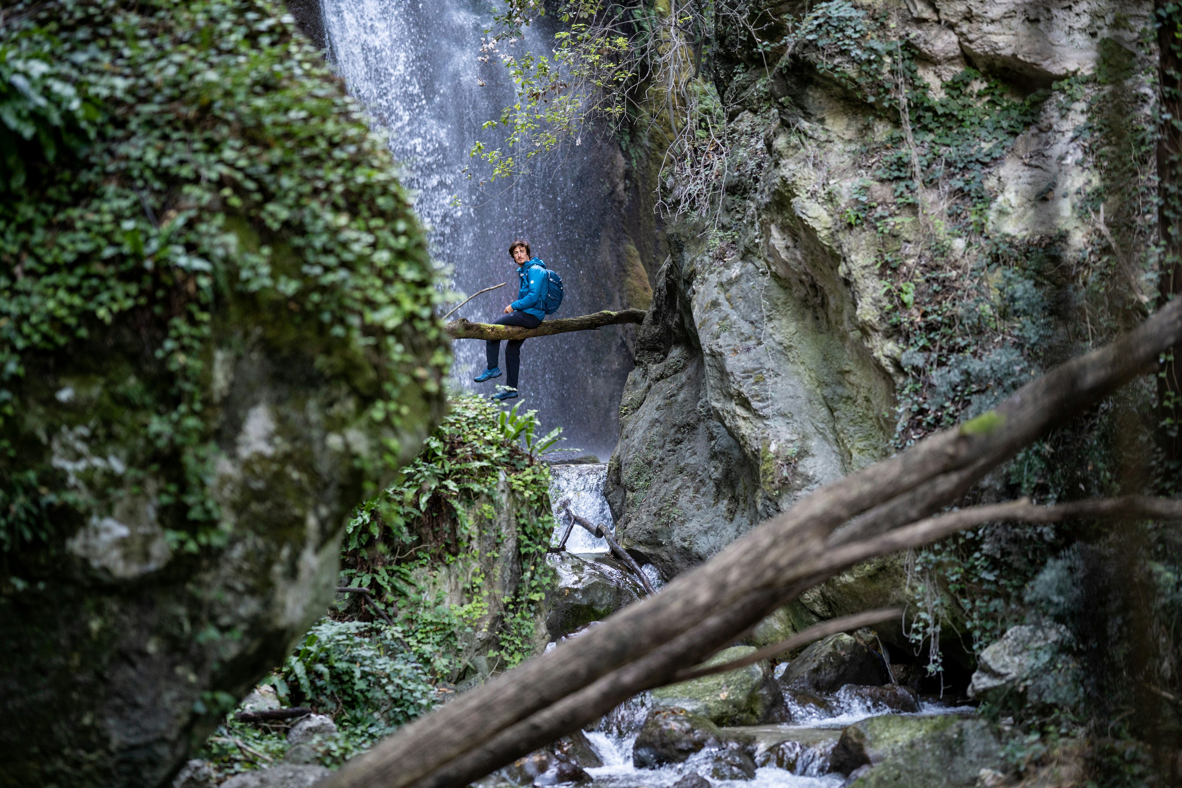Mann sitzt auf einem Baumstamm vor einem Wasserfall.