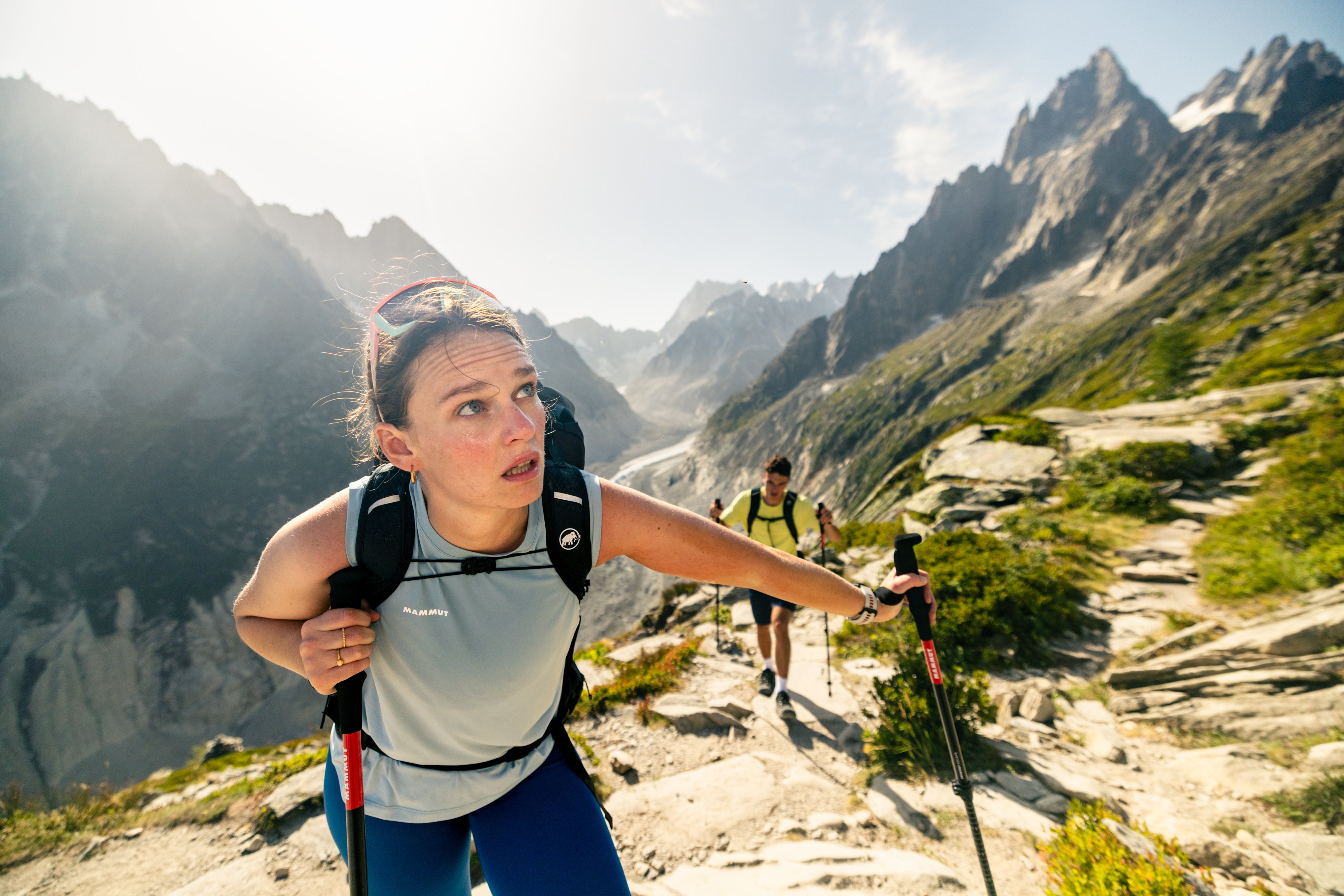 Woman wearing Mammut gear hikes uphill on a rocky mountain trail with trekking poles, another hiker follows behind, sunny day.