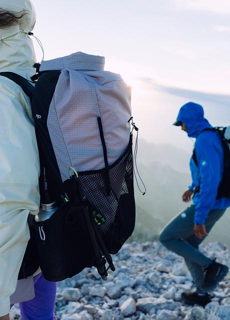 Two hikers wearing Mammut hooded jackets and backpacks trek across rocky mountain terrain at sunrise, showcasing premium outdoor gear for mountaineering.