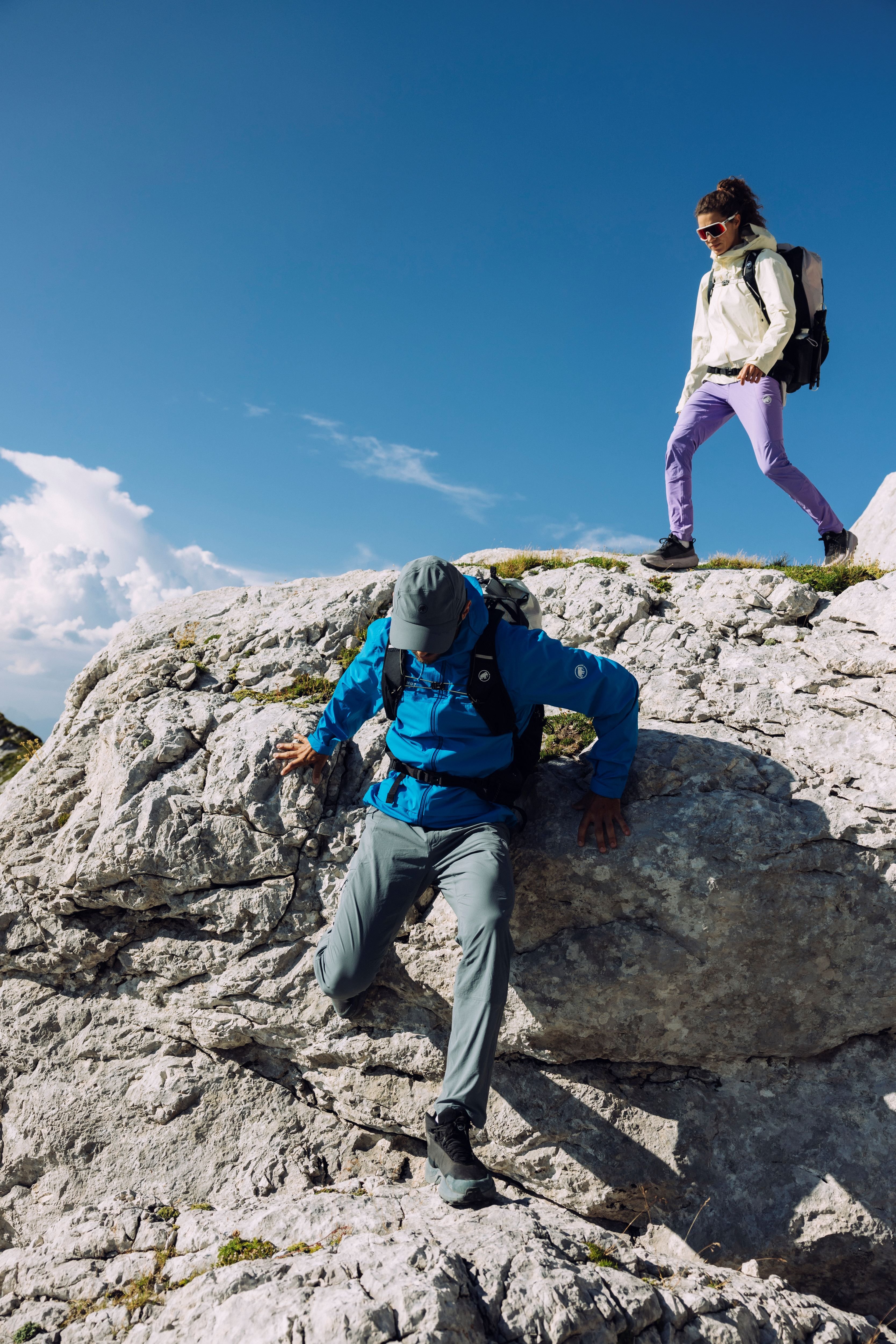 Two hikers wearing Mammut gear navigate a challenging rocky terrain under a clear blue sky. One adventurer descends a rock formation while the other closely follows.