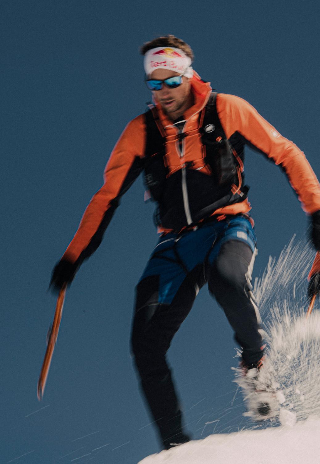 Mammut climber in an orange jacket and black pants with ice axes, tackling a snowy slope against a clear sky.