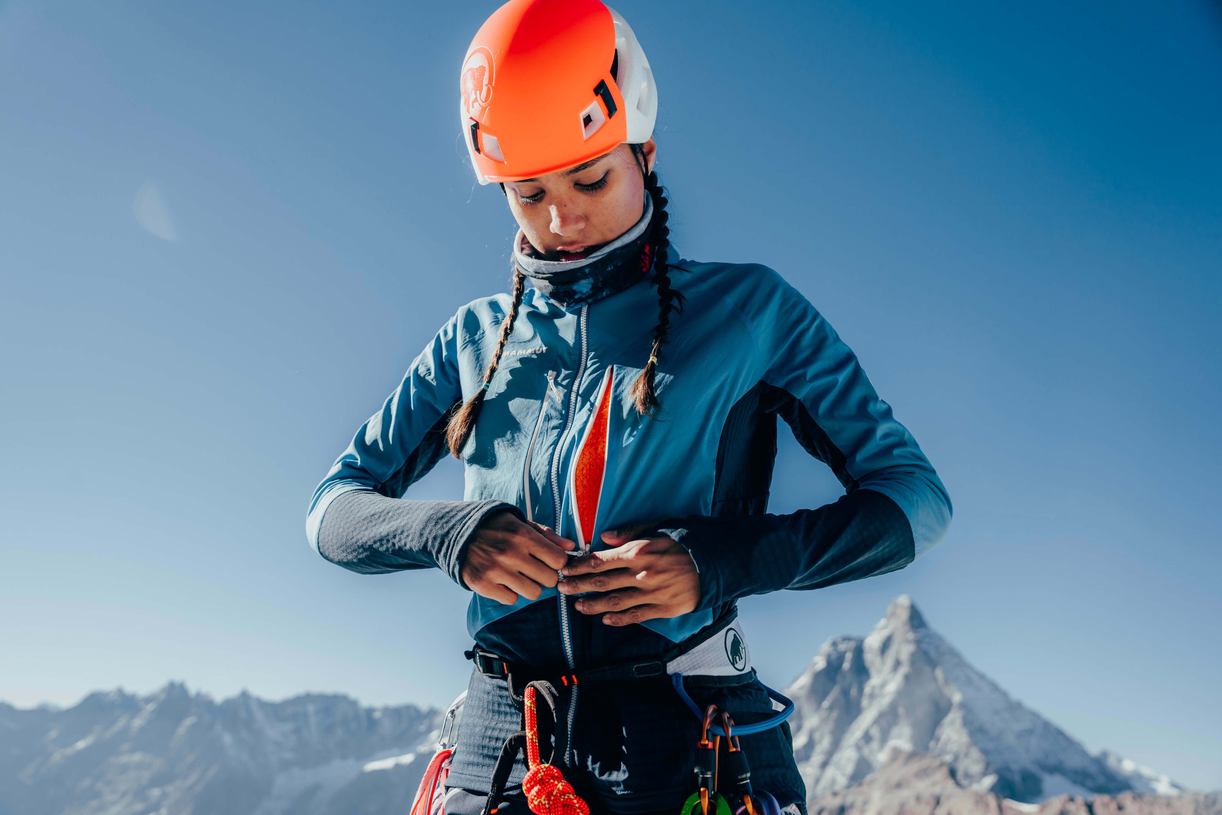 A mountain climber wearing a blue Mammut jacket and an orange helmet is adjusting their climbing gear. A snowy peak rises dramatically in the background.