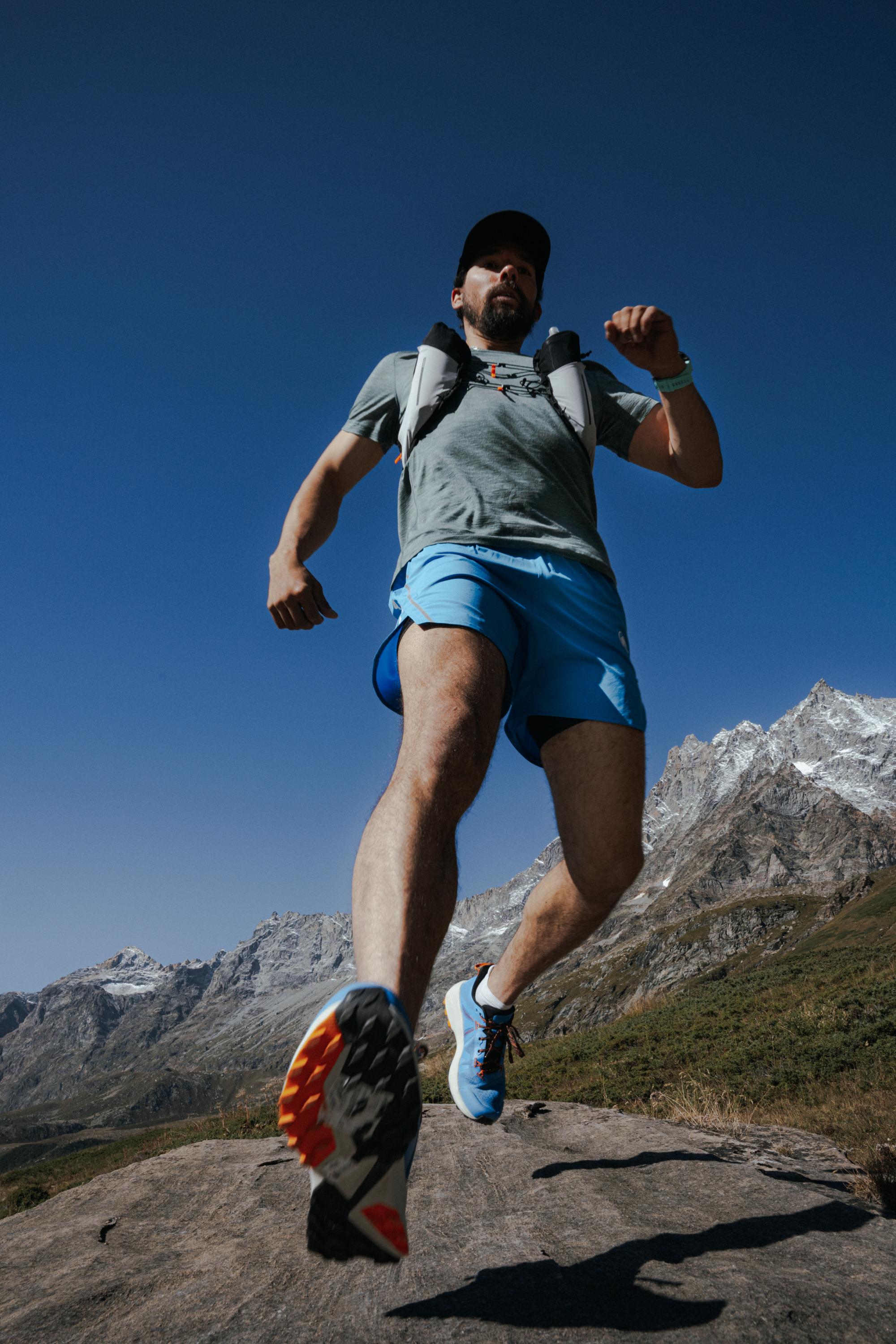 Low-angle shot of a person running on rugged terrain wearing Mammut gear, with majestic mountains in the background under a clear blue sky.