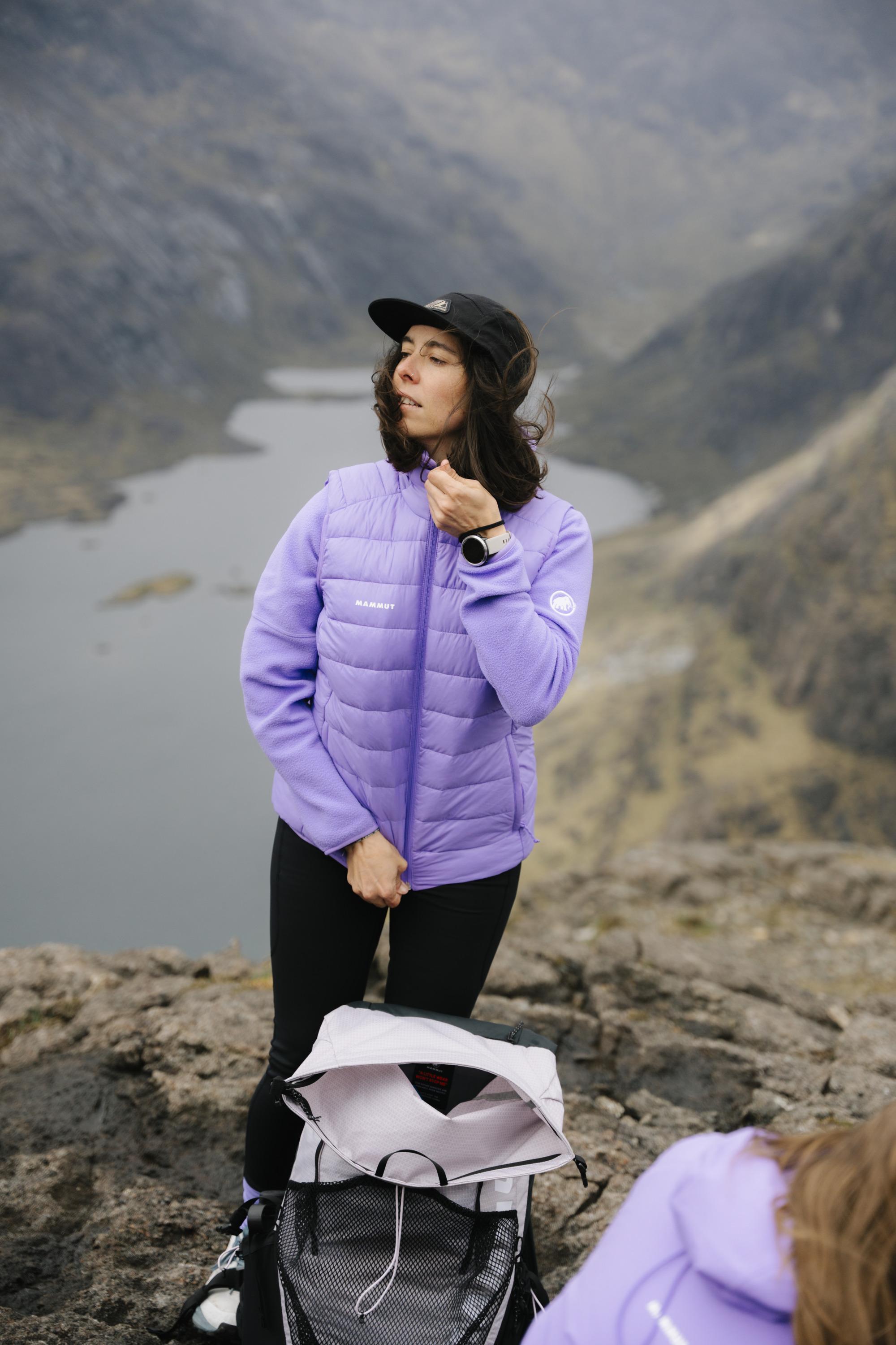 Woman in a Mammut light jacket sits outdoors, pulling the collar over her mouth, looking thoughtfully to the side.