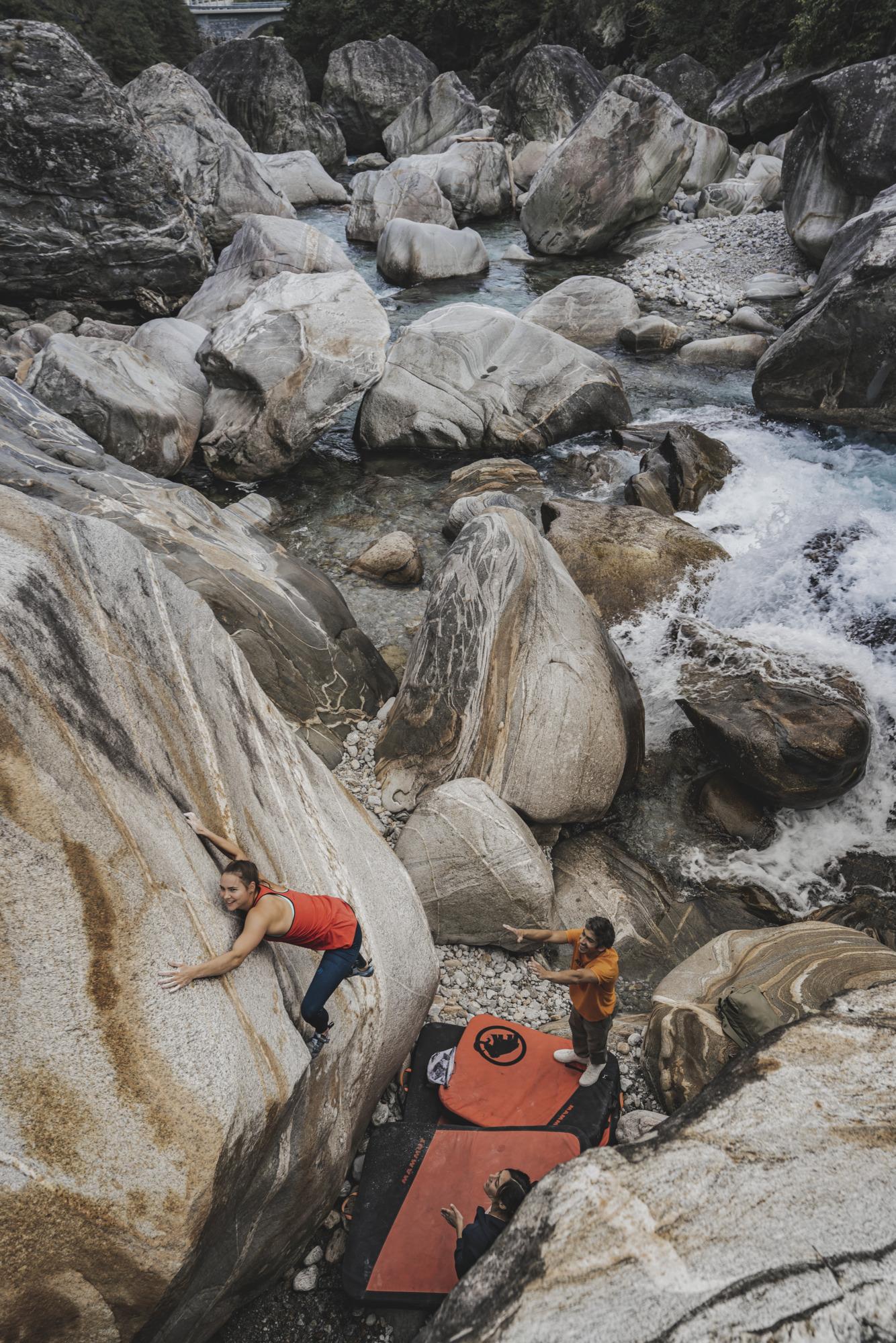 A climber in a red Mammut tank top scales a massive boulder near a riverside, while two fellow climbers stand below on red Mammut crash pads providing support.