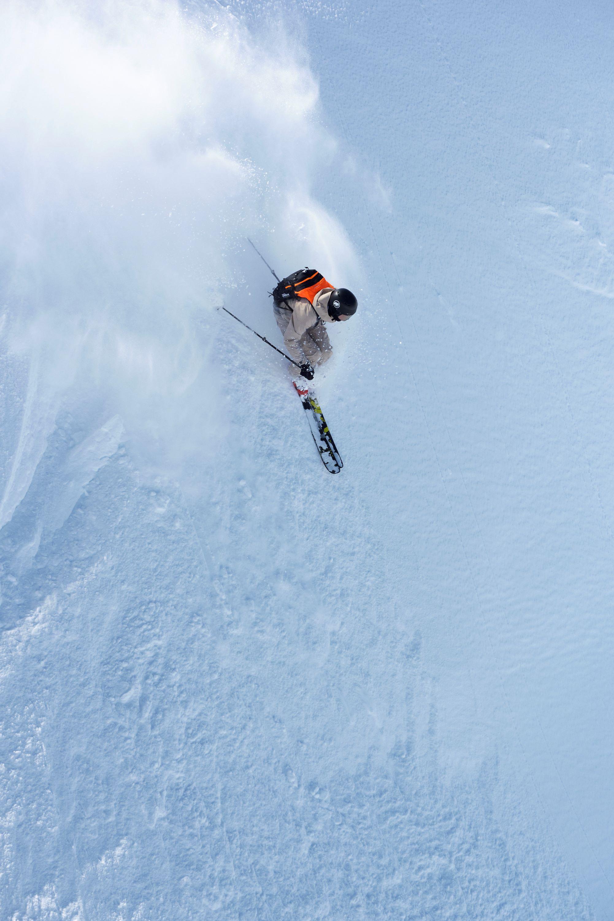 Skier in mid-air performing a jump, surrounded by snow spray on a steep, snowy slope, wearing Mammut gear.