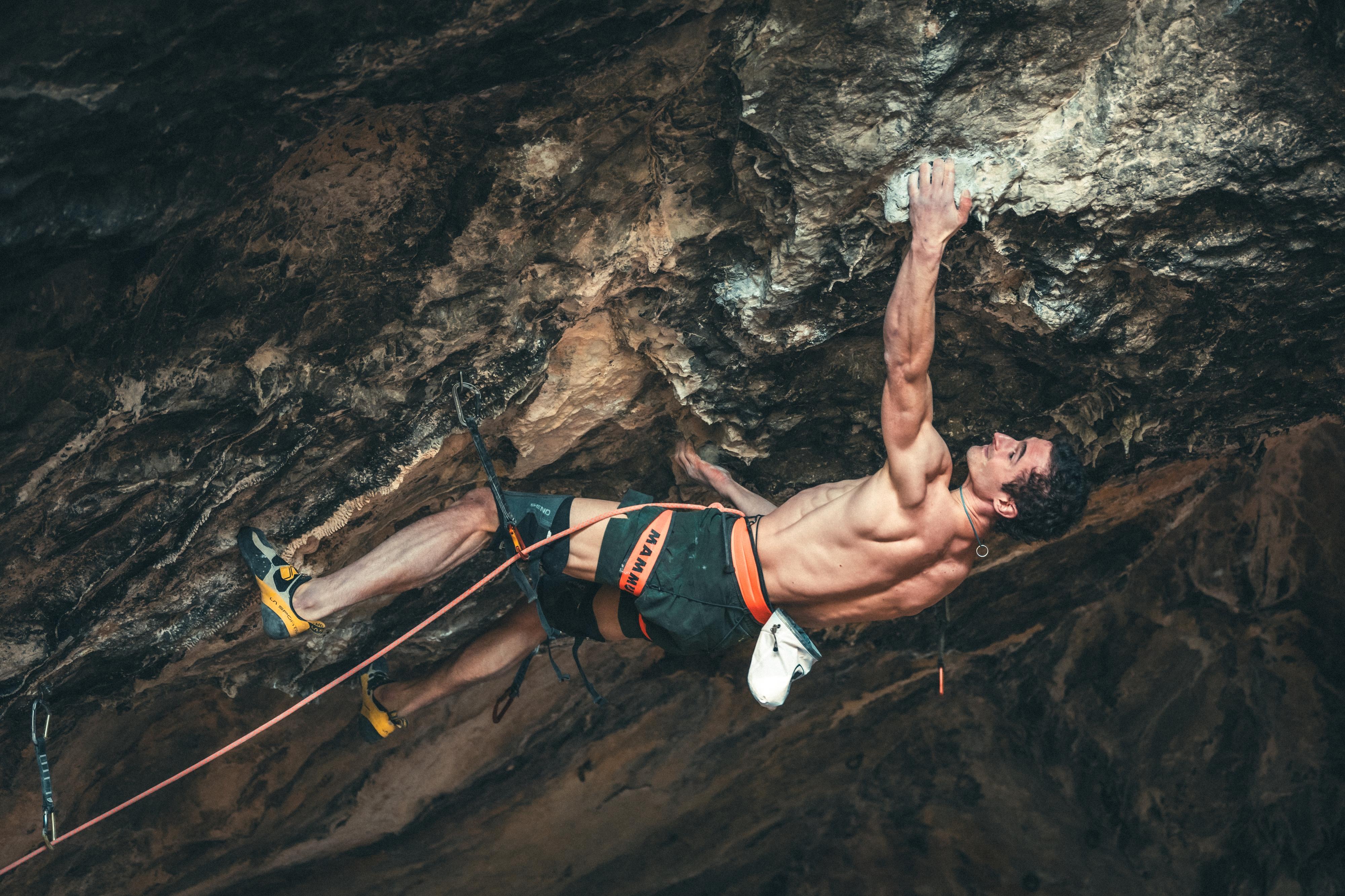 A Mammut-equipped rock climber ascends a challenging, overhanging route in a cave, using high-performance gear and demonstrating significant muscular effort.
