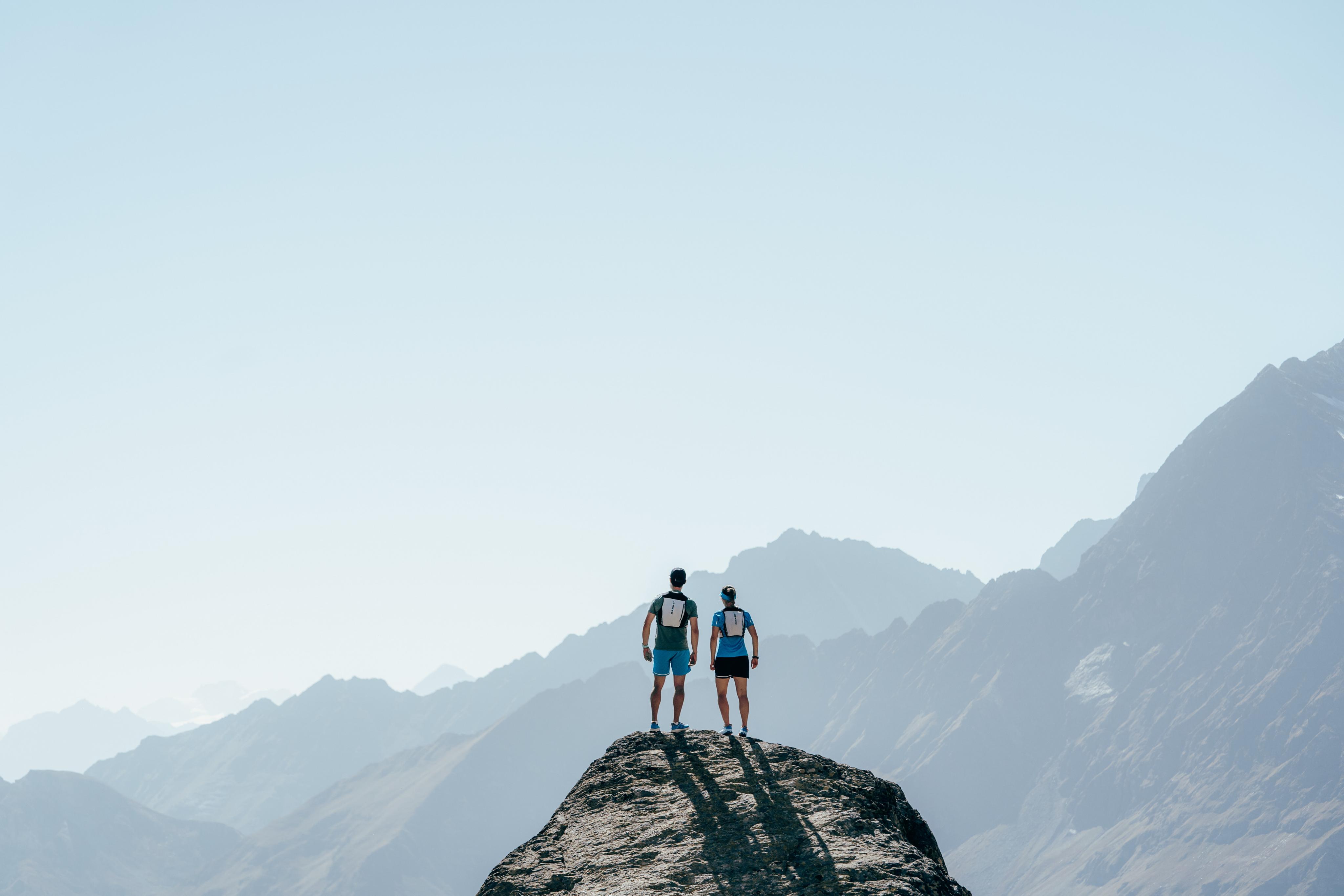 Two mountaineers in Mammut gear standing on a mountain peak, admiring vast alpine ranges under a clear blue sky.