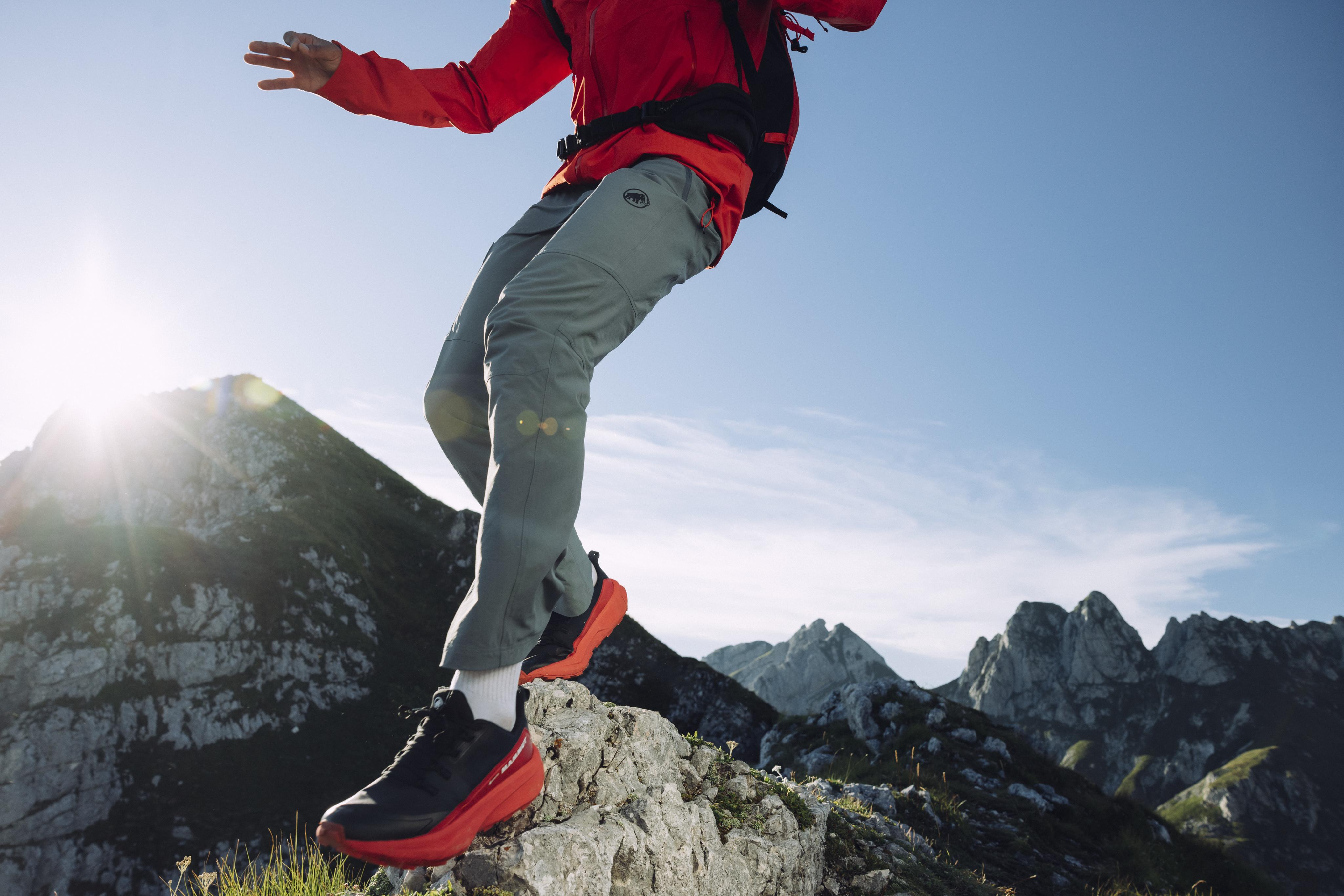 Hiker in Mammut gear navigating a rocky mountain trail with a sunny Alpine backdrop.