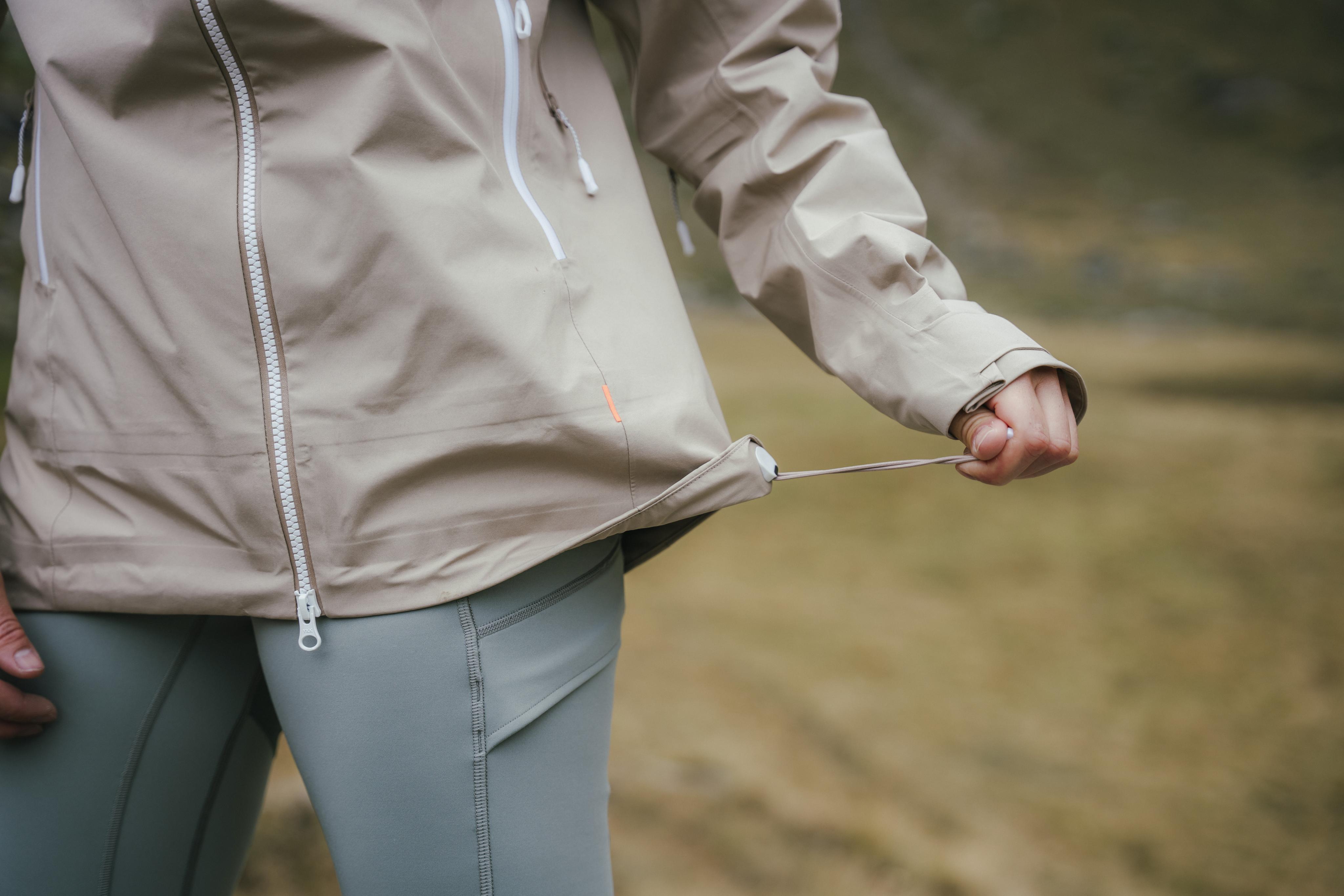 Person wearing a Mammut tan jacket adjusts the under-sleeve zipper while standing in a mountainous area outdoors.