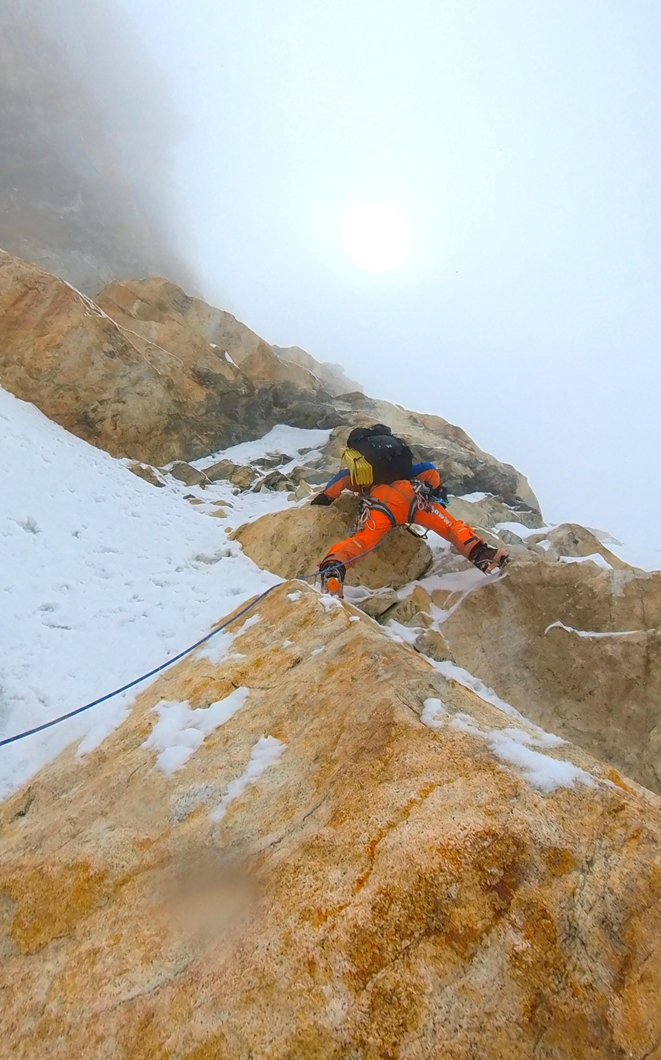A climber in Mammut's bright orange gear scales a snowy, rocky mountain slope under a cloudy, foggy sky.