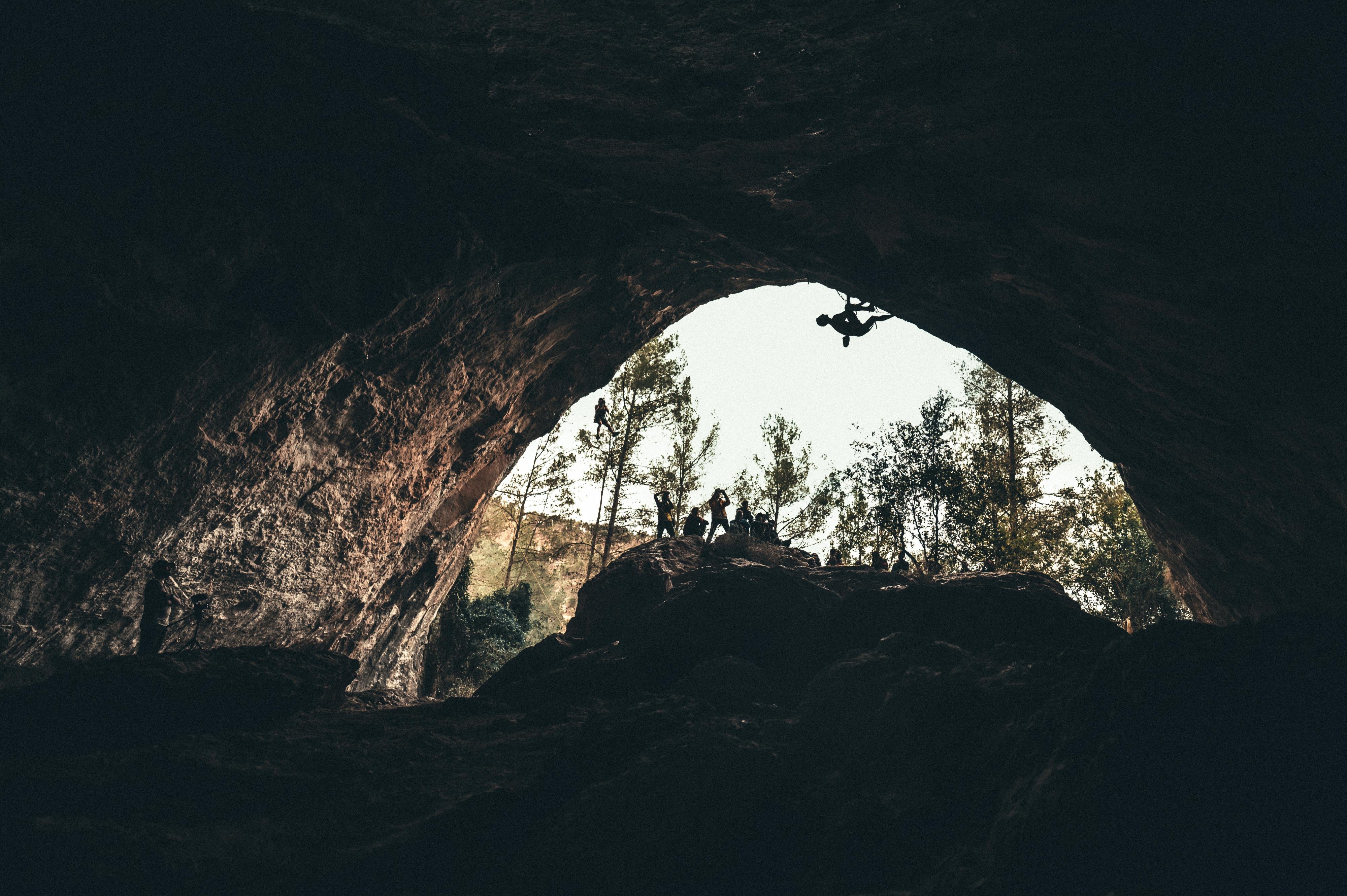A climber ascends inside a large cave wearing Mammut gear, with onlookers in the background and trees visible through the cave's opening.