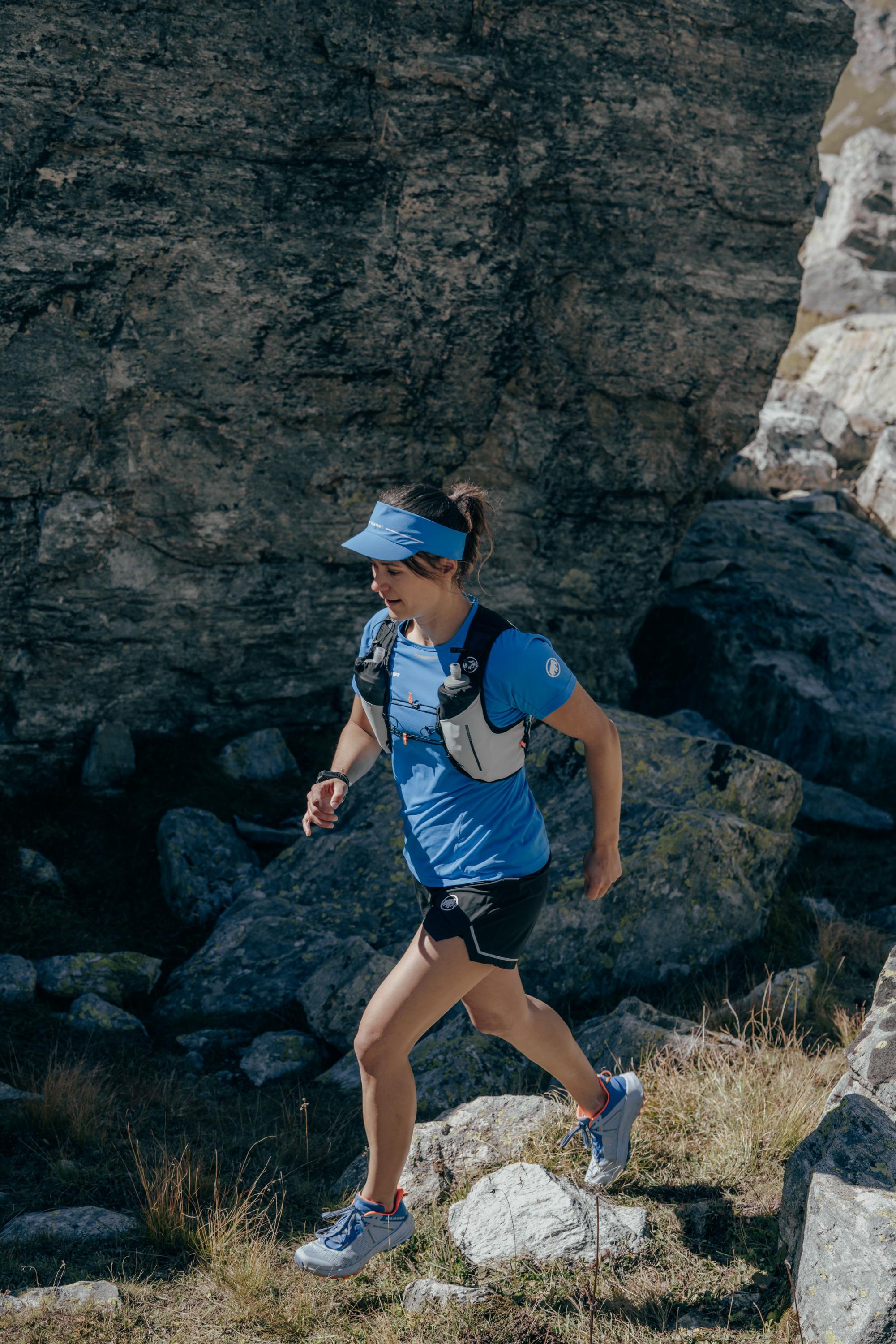 A trail runner in Mammut athletic wear and a visor navigates a rocky path beneath a towering boulder.