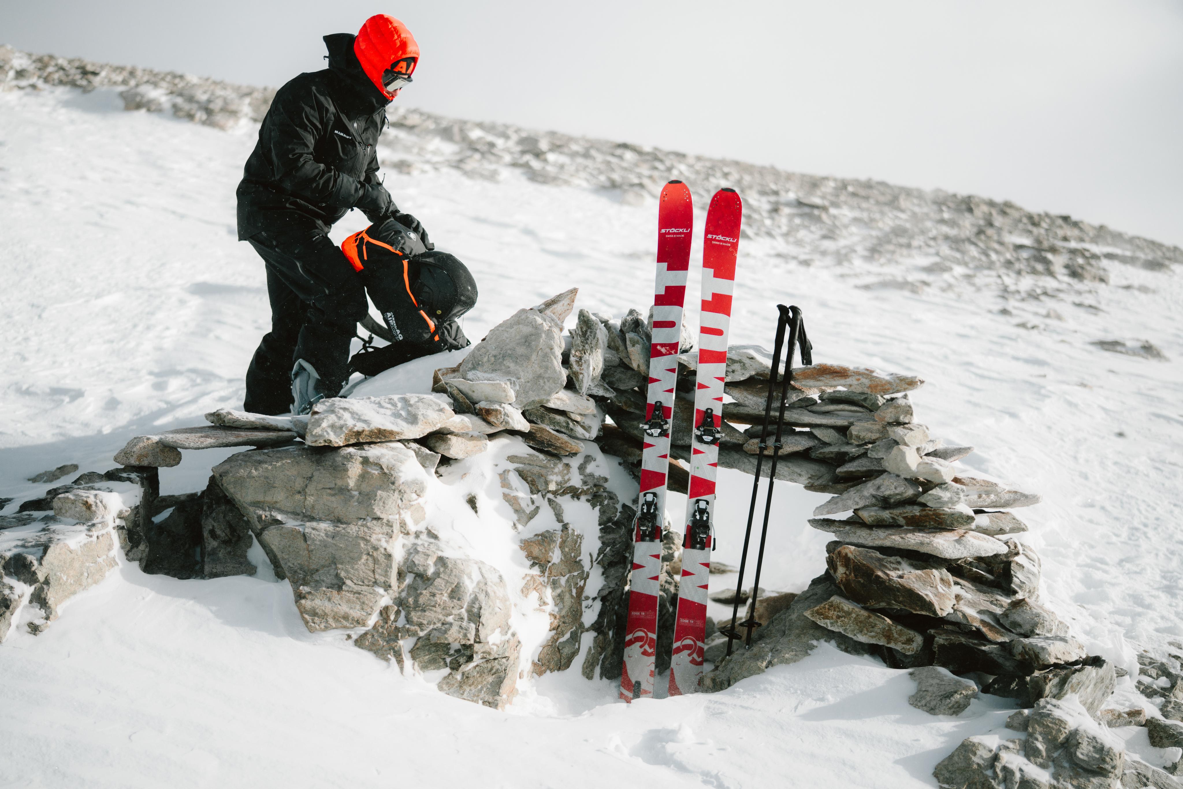 A skier in a yellow jacket, wearing Mammut gear, zips up his coat on a snowy mountain with evergreen trees and rugged peaks in the background.