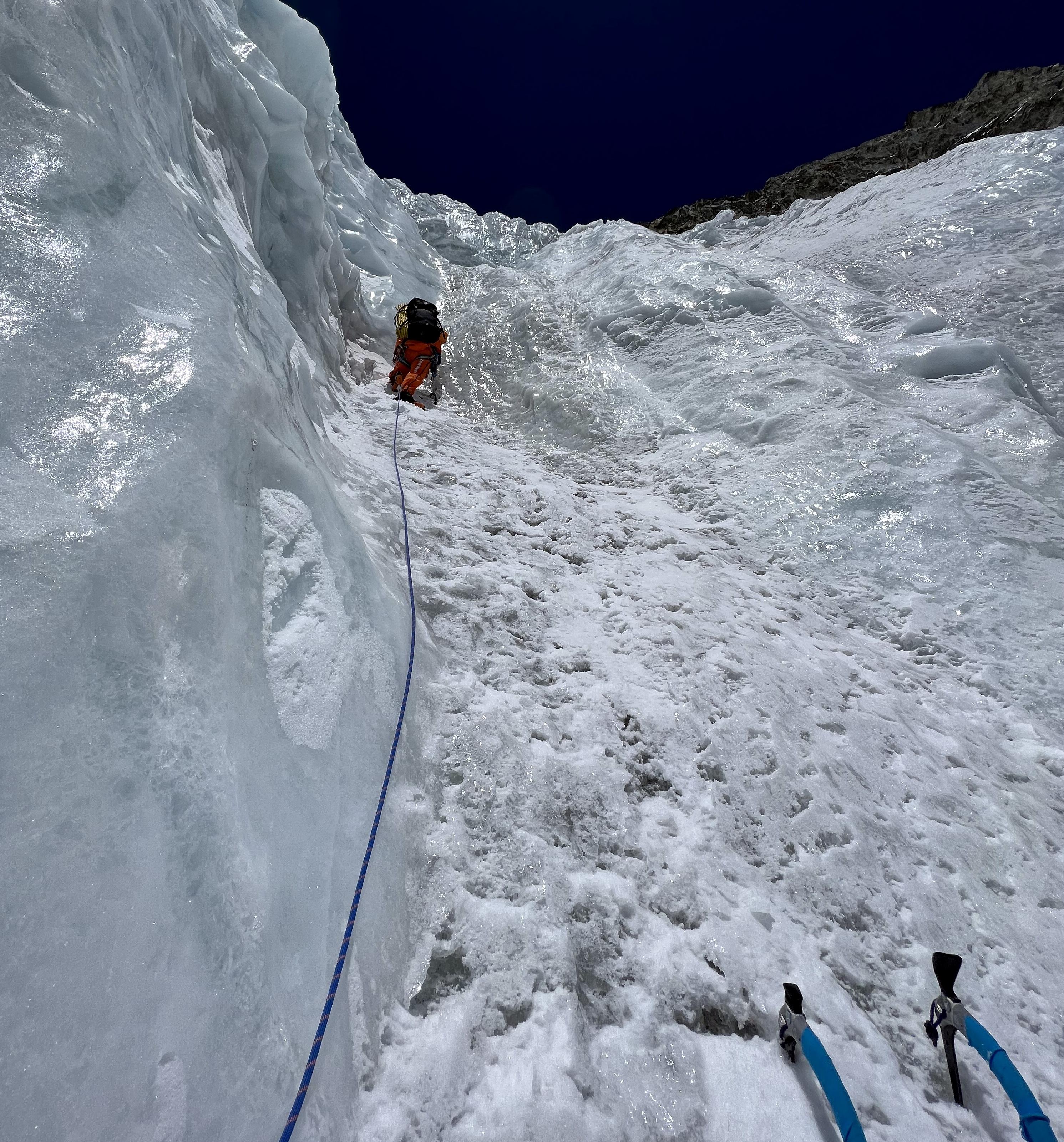 A climber wearing Mammut gear ascends a steep, icy slope using a rope and ice tools, with a deep blue sky above.