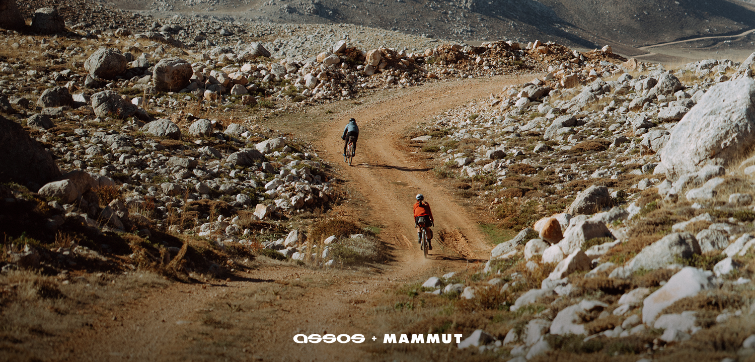 Cyclist wearing a Mammut-branded helmet rides along a gravel path in a rocky, dry mountain landscape.